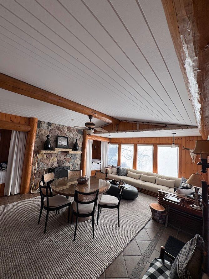 A rustic living and dining room with a large stone fireplace, exposed log beams, a white paneled ceiling, and rug.