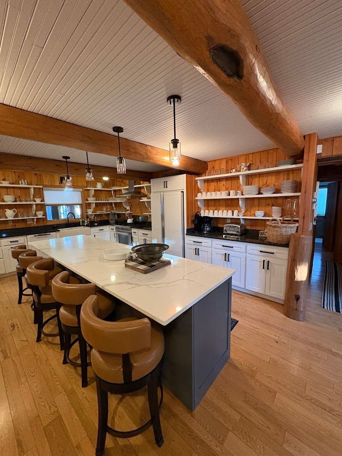 A rustic kitchen with wood-paneled walls, a large white-topped island with tan bar stools, and open white shelving.
