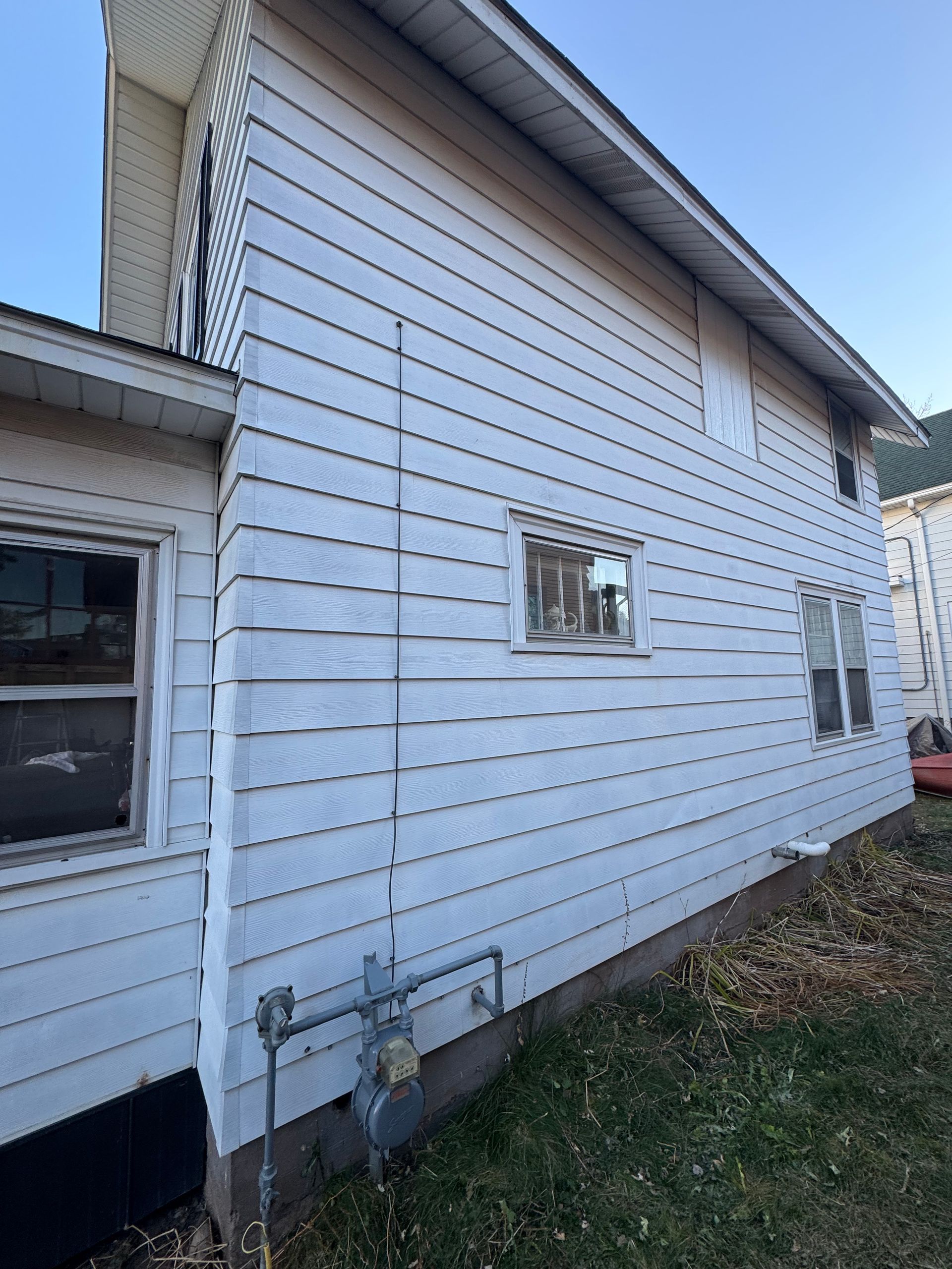 A side view of a white, two-story house with horizontal siding, two windows, and an outdoor gas meter near the ground.