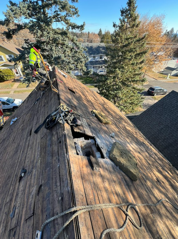 A worker in a high-visibility jacket stands on a roof showing a large hole in the wooden decking.