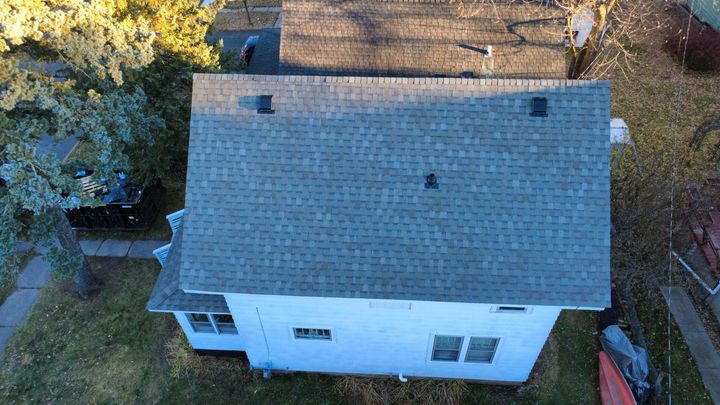 An aerial, slightly angled view of a house with a gray shingled roof and white exterior walls in a residential yard.