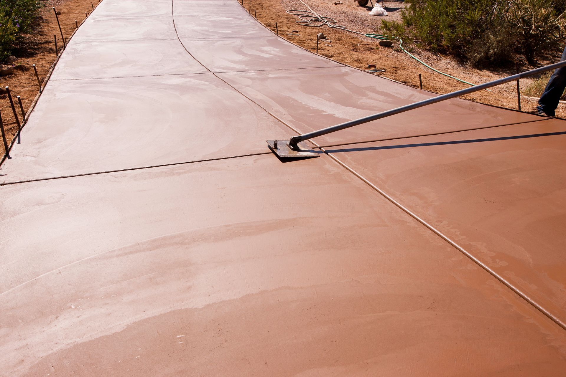 Freshly poured concrete driveway with a metal cable brace across it, beside dirt and rocks