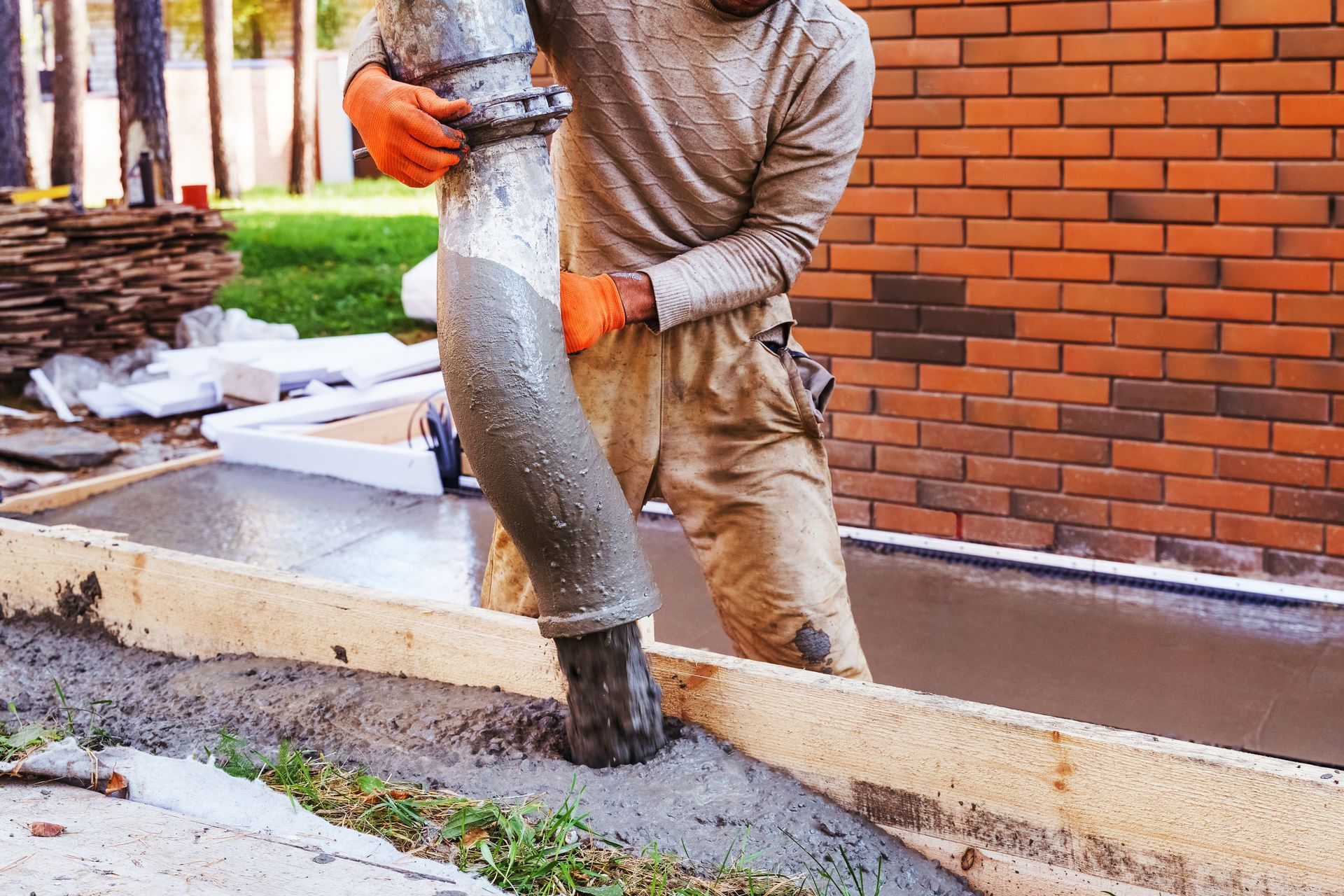 Worker pours concrete from a hose into a form. Orange gloves, brick wall in background.