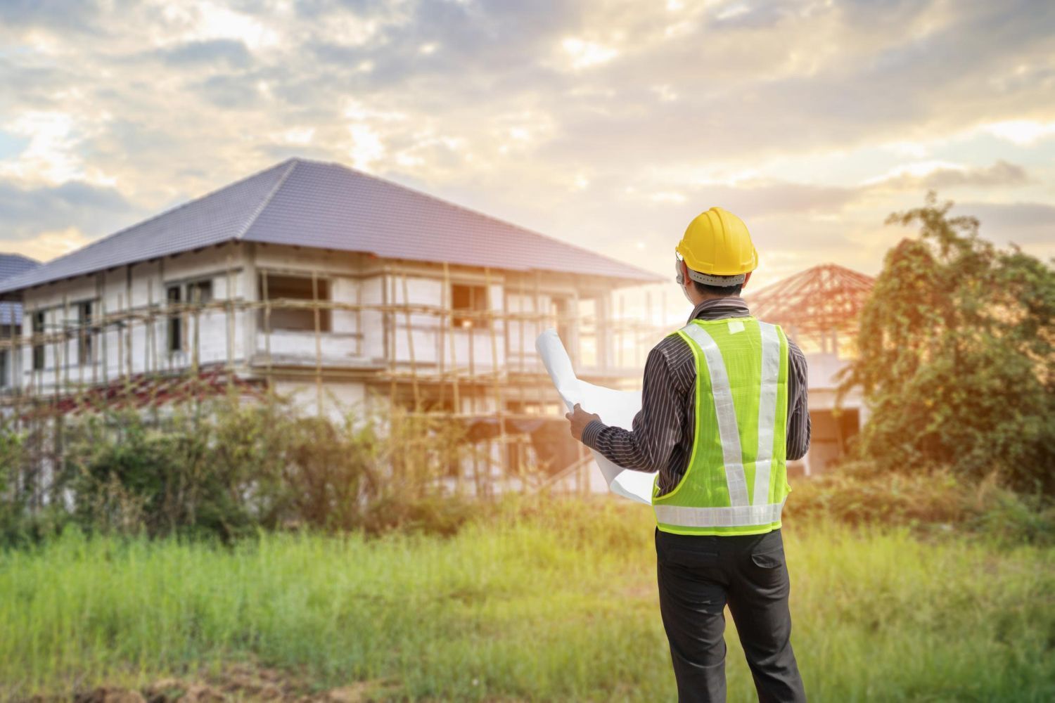 Construction worker in a yellow hard hat and safety vest, reviewing blueprints on a building site.