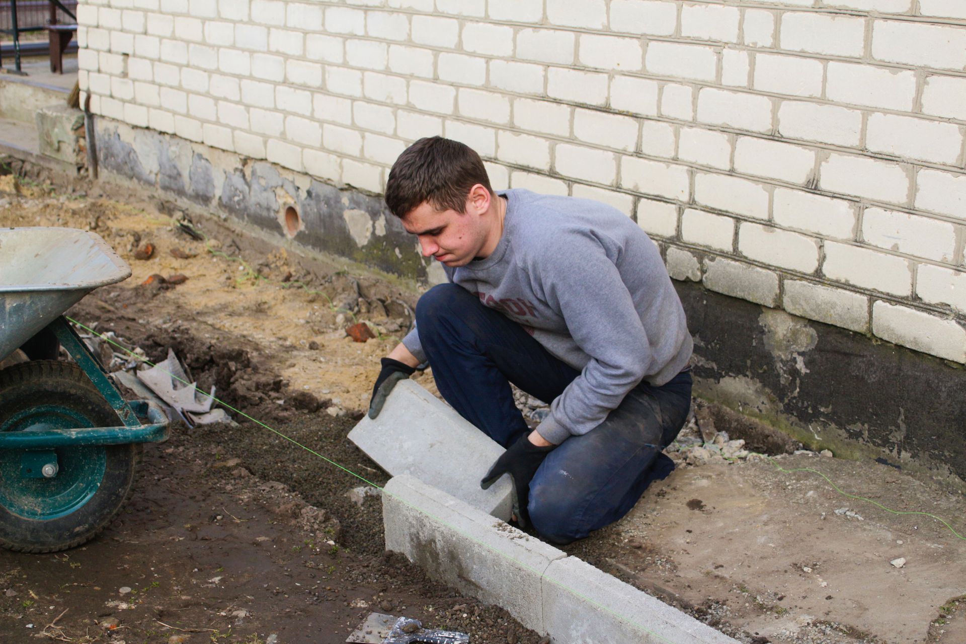 A man is kneeling down next to a wheelbarrow while working on a sidewalk.