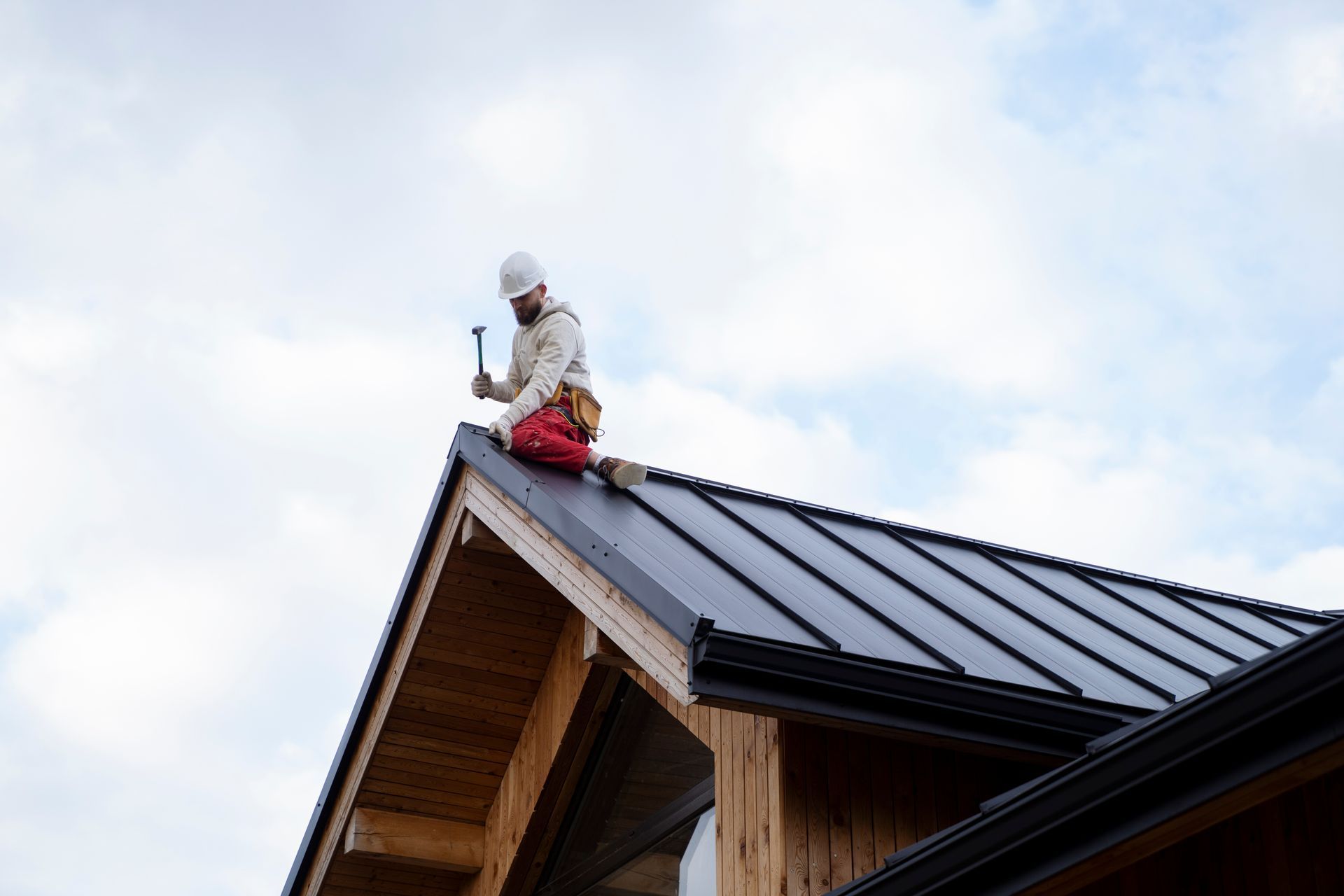 Worker on a roof near the ridge, installing black metal roofing under a cloudy sky