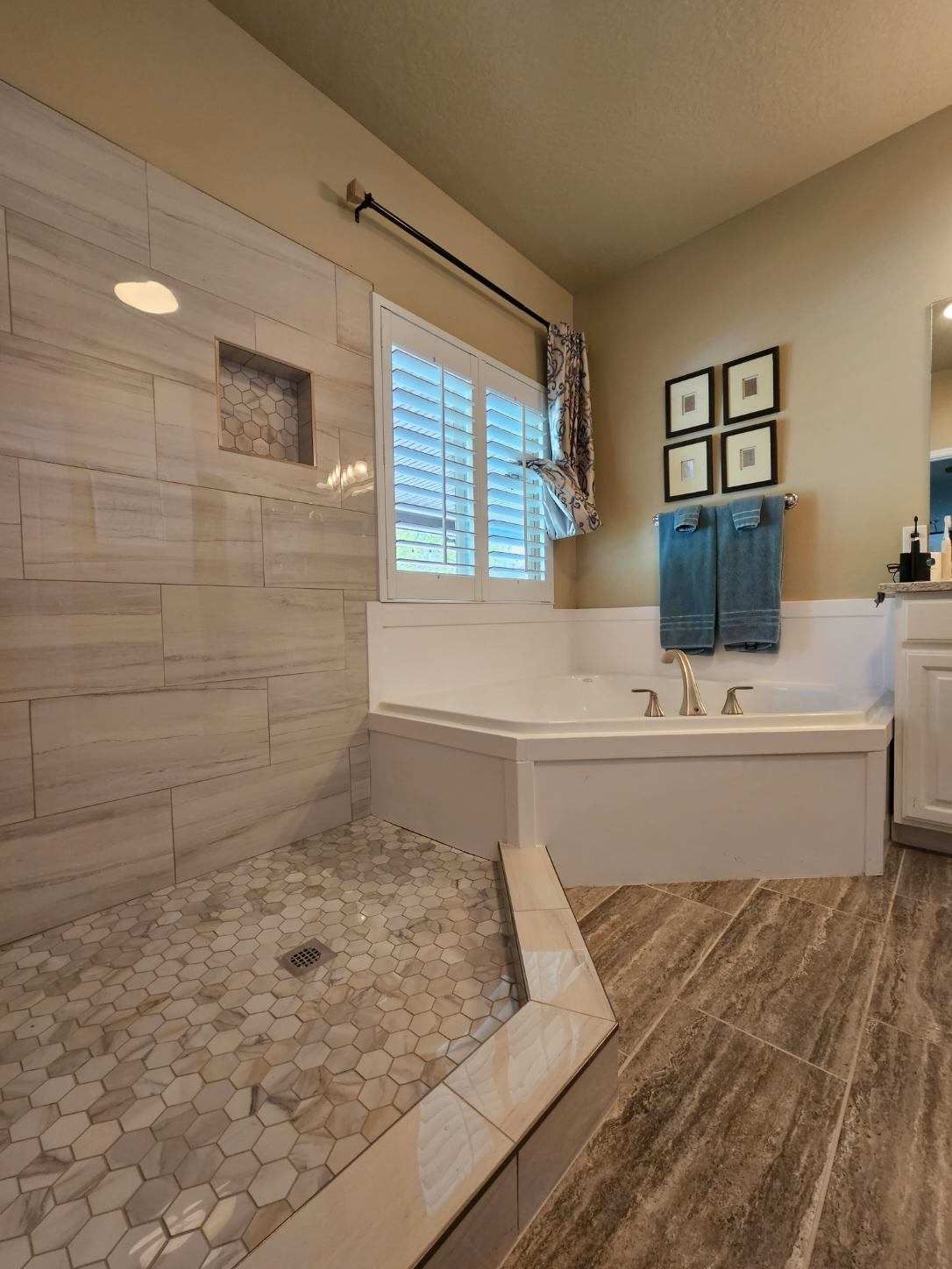 Bathroom with a walk-in shower, built-in tub, and shuttered window. Beige walls, gray tile, and blue towels.