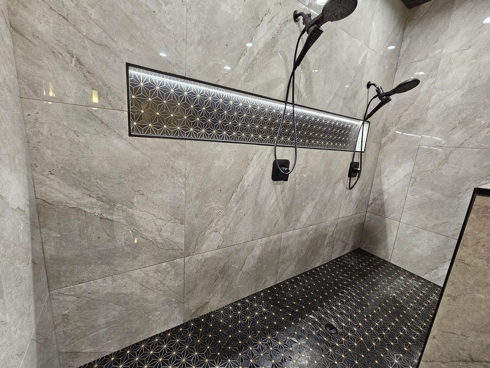 Modern shower with gray marble walls and dark mosaic floor, with overhead shower heads and long light fixture.