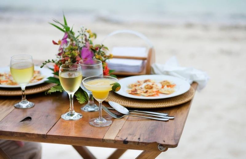 A wooden table with plates of food and wine glasses on it on the beach.
