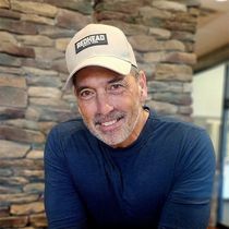 Man in a navy shirt and beige cap smiling indoors against a stone wall.