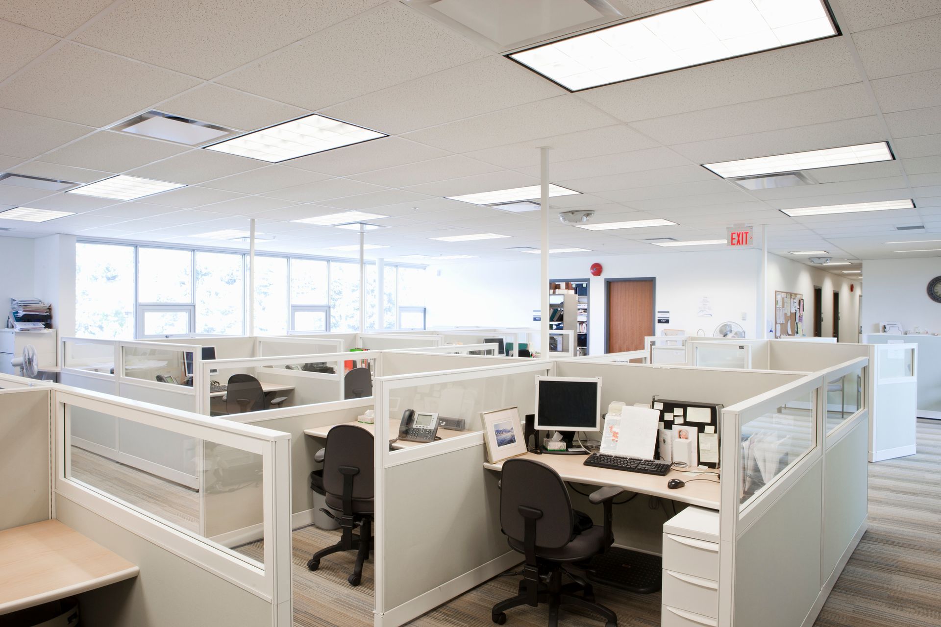 Office cubicles in a bright, open space. Beige walls, desks, and chairs. Large windows.