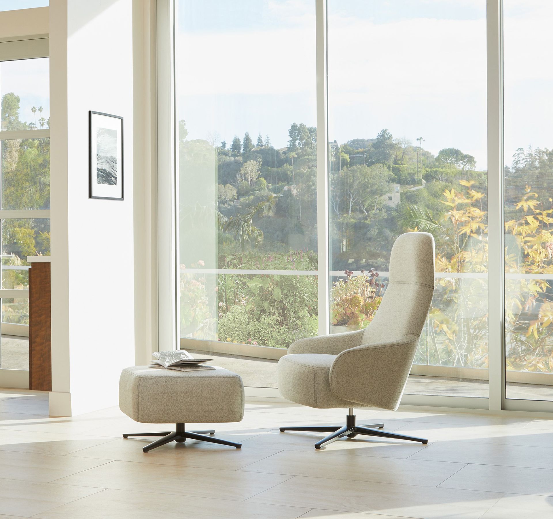 A beige chair and ottoman with a swivel base, positioned near a large window overlooking greenery.