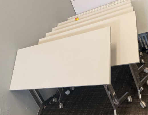 Stack of white rectangular tables with silver legs, in a room.