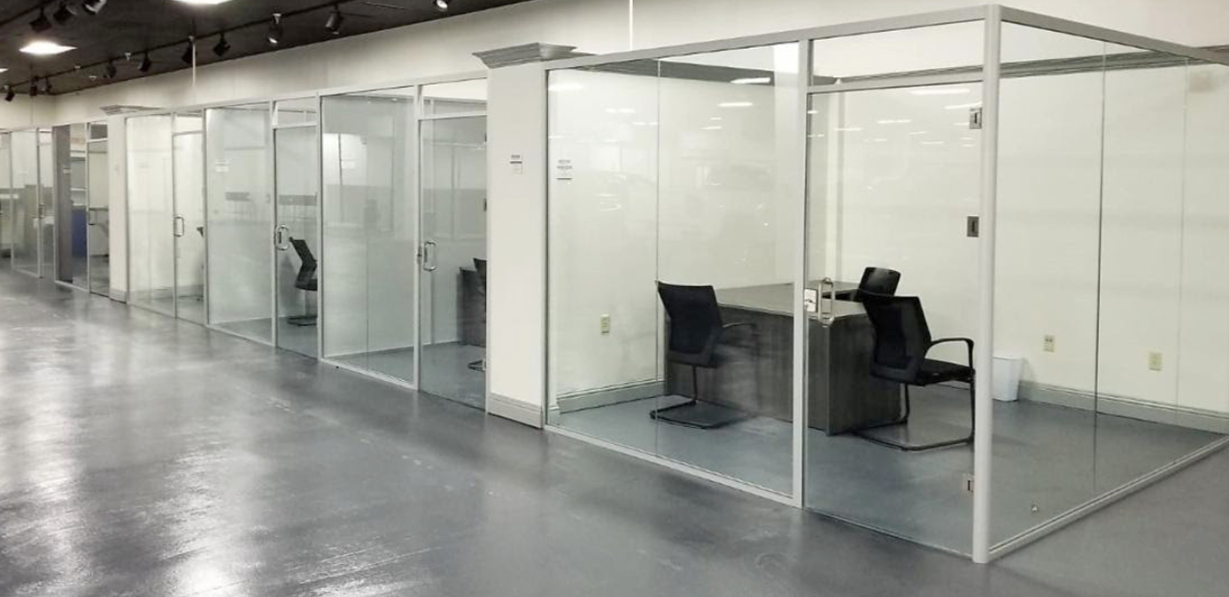 An office corridor with several glass-walled cubicles. Each cubicle has a desk and chairs.