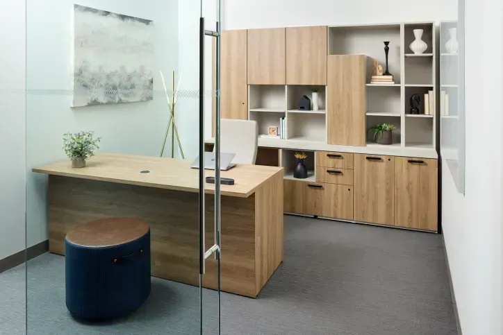 A modern office with wooden desk, storage, and blue stool. Glass door. Light wood and gray tones.