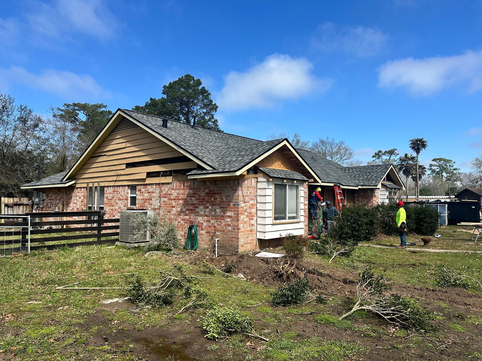 Brick house with partial roof repair, workers in safety vests, blue sky, and overgrown yard.