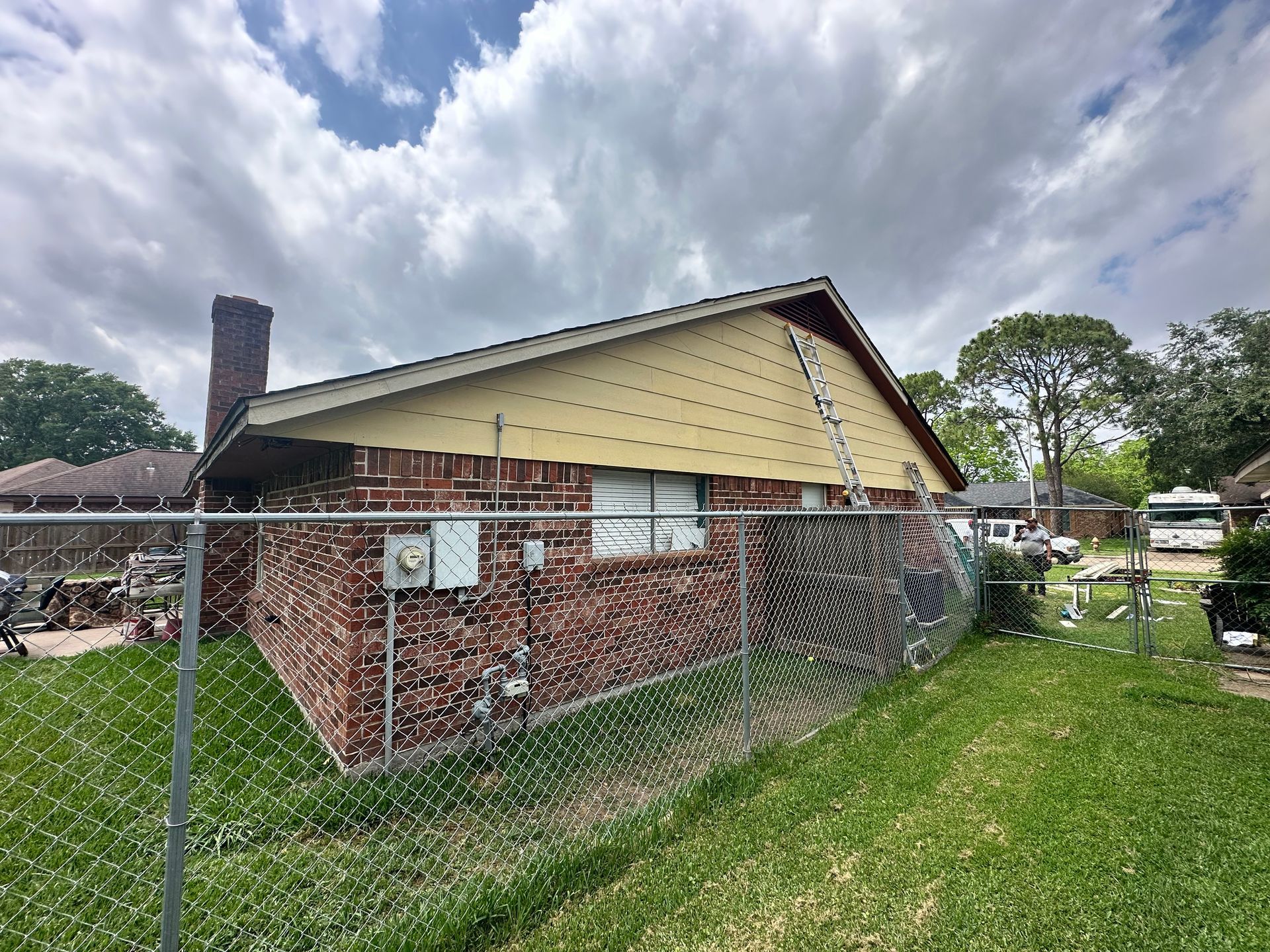 Brick house with a chain-link fence in a grassy yard, under a cloudy sky.