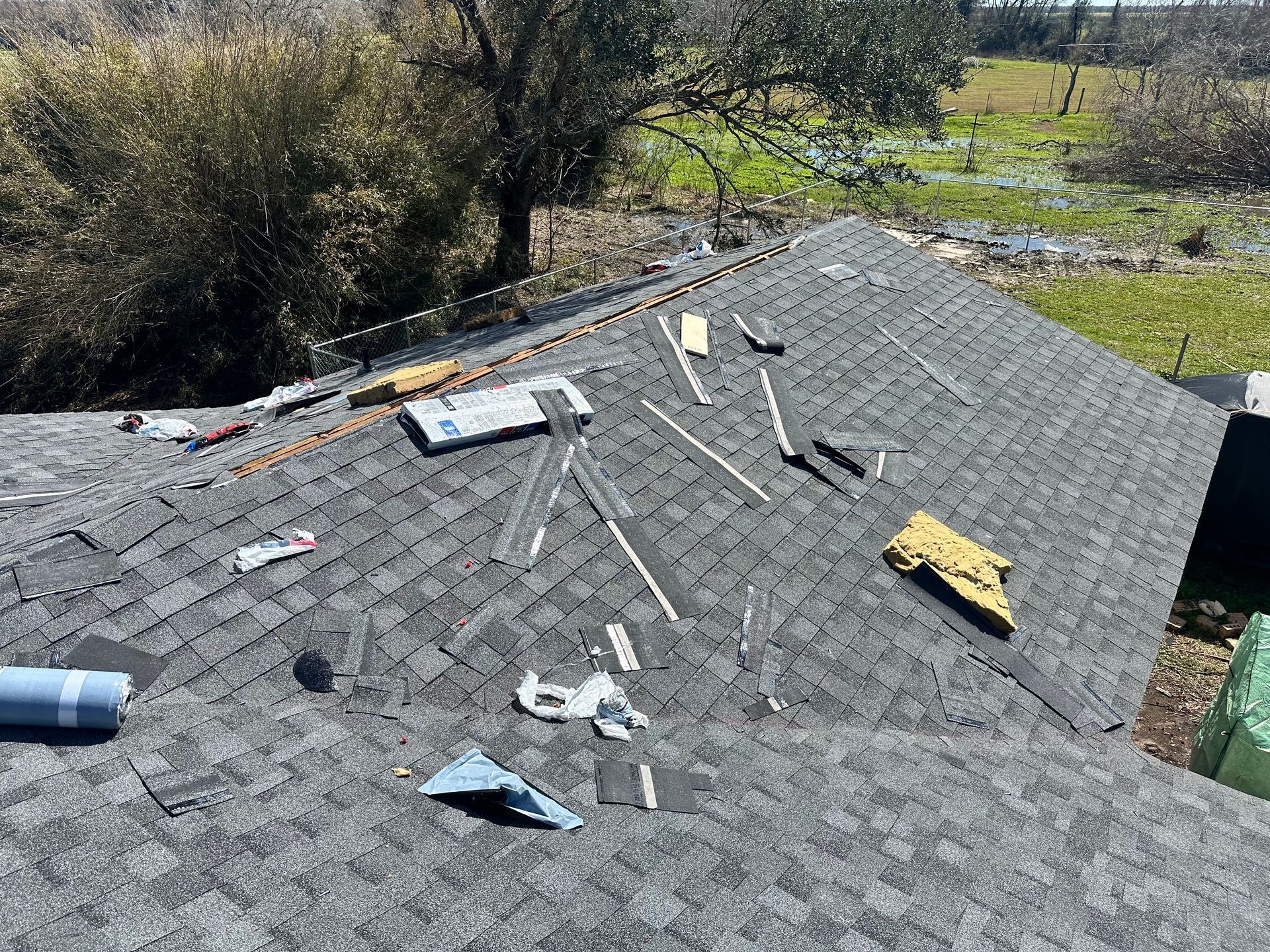 Damaged asphalt shingle roof with debris scattered. Blue sky background, trees in distance.