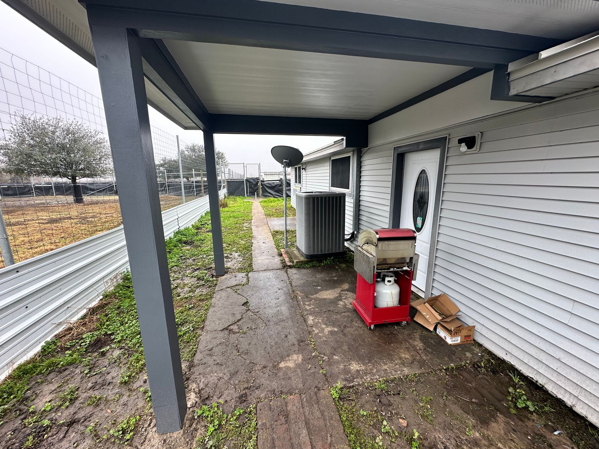 Covered walkway next to a white house, concrete path, gray posts, red machine, grass and fence.