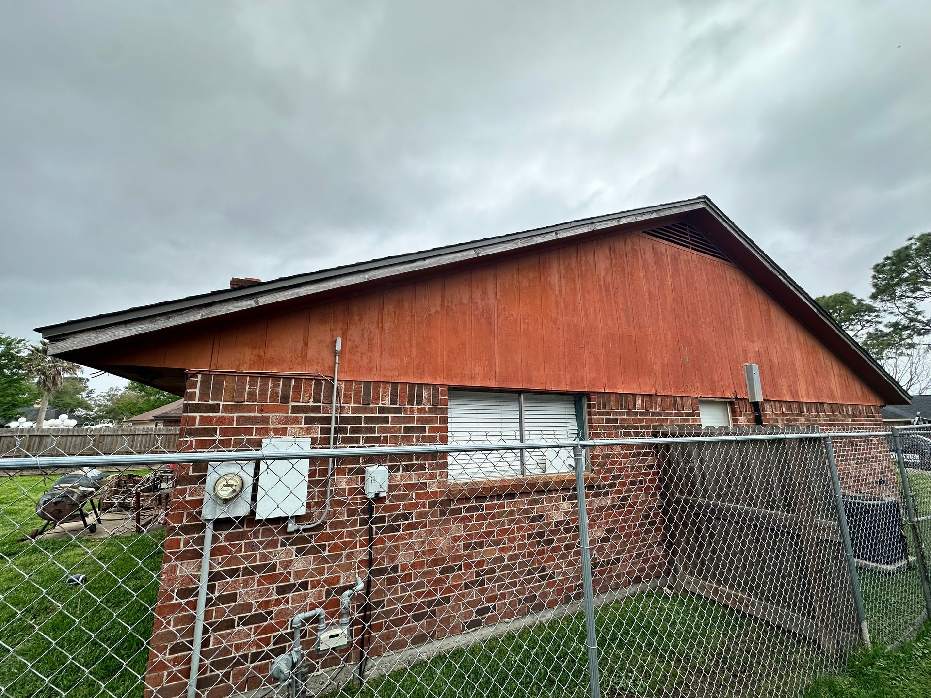 Brick house with red wooden siding, behind a chain-link fence, under a cloudy sky.