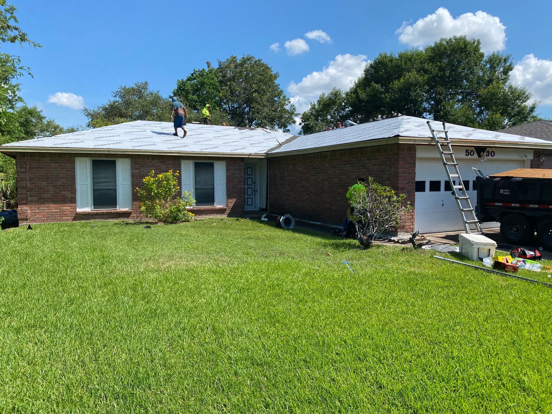 Workers replacing a roof on a brick house; ladder leans against the garage.