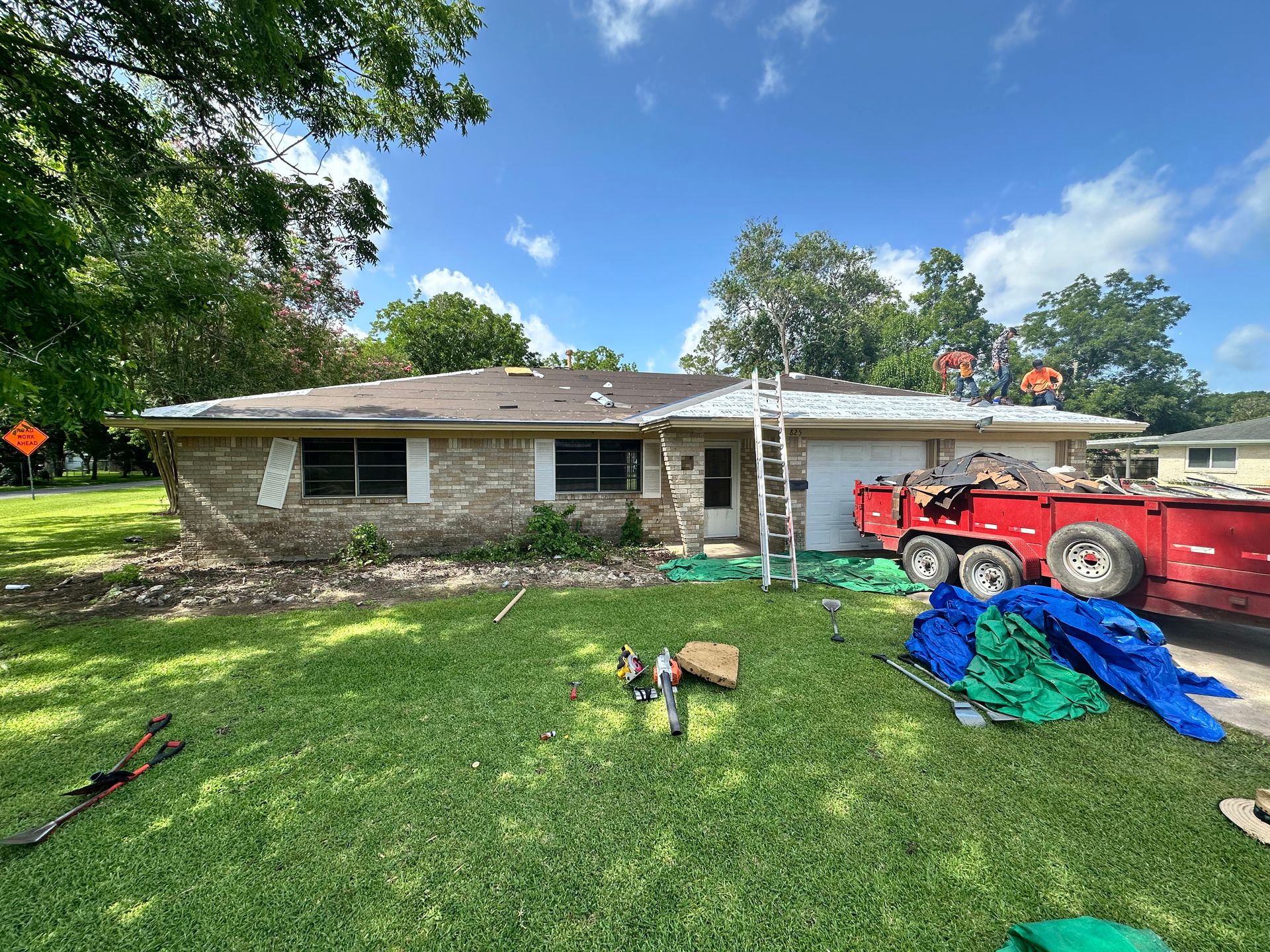 House with a partially removed roof; a ladder, truck, and tarps are on the lawn.