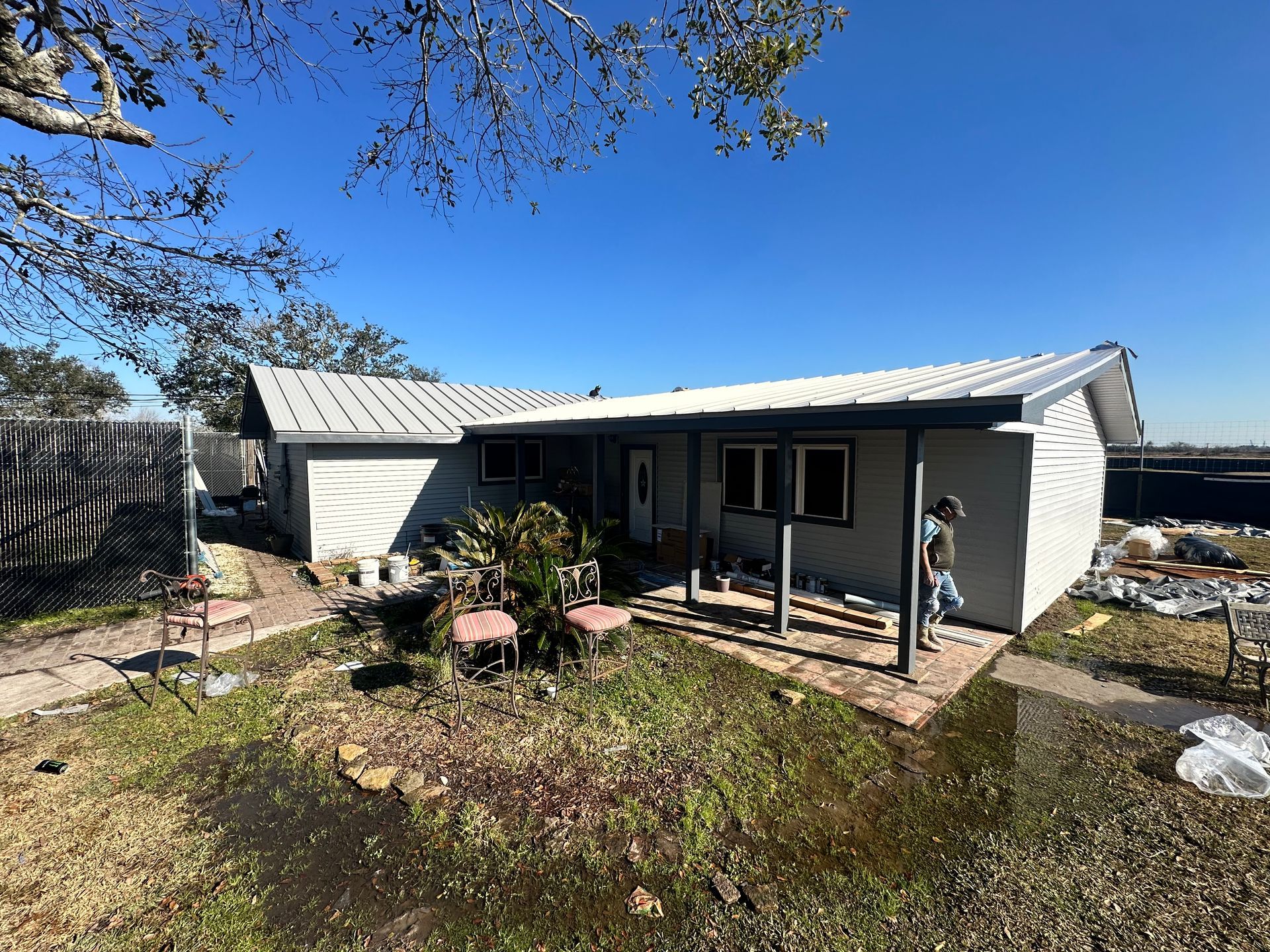 A gray house with a metal roof under a bright blue sky. Front yard has plants and two chairs.