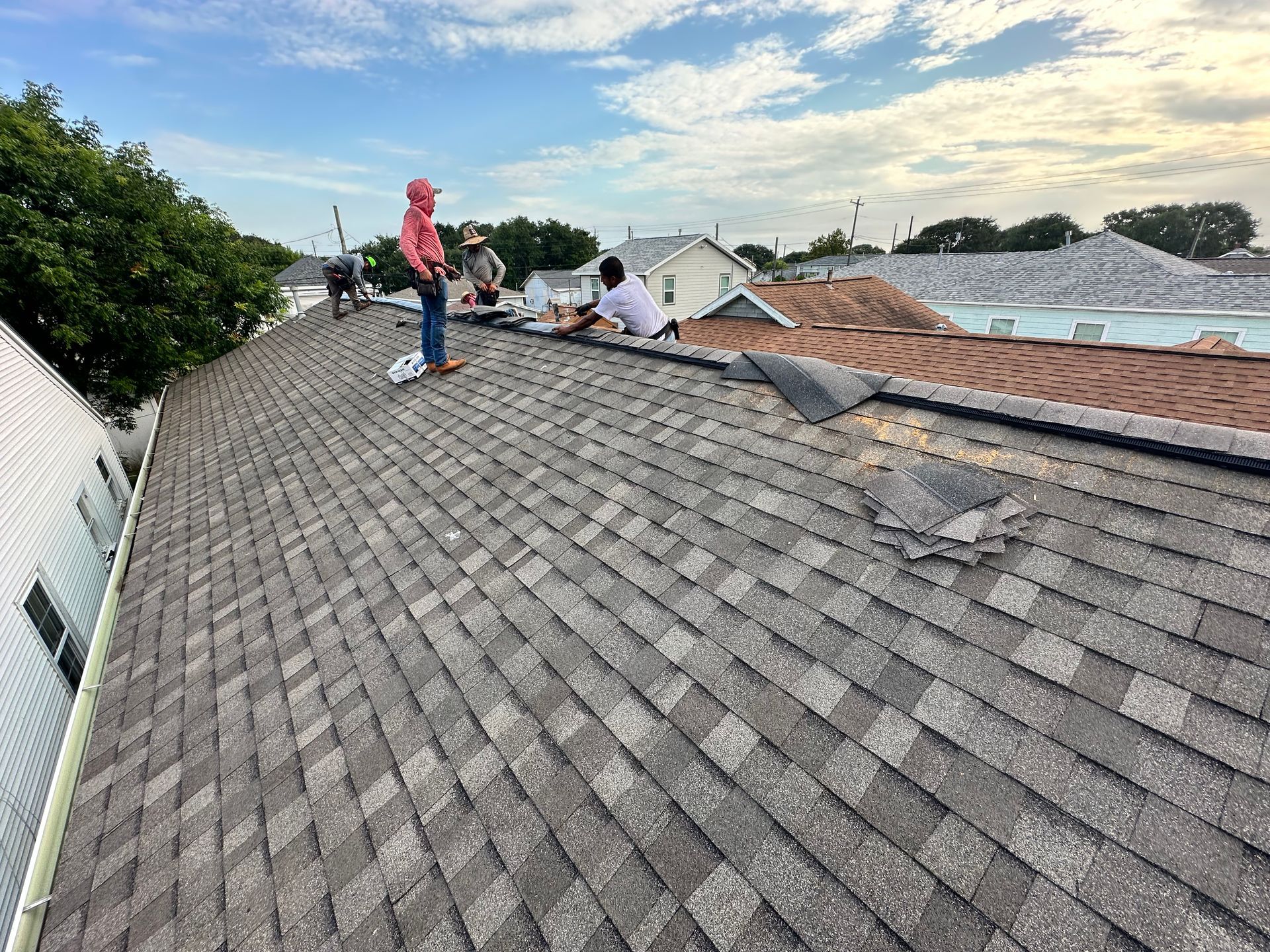 Roofers repairing a damaged shingle roof. Gray shingles, blue sky, and other houses are in the background.