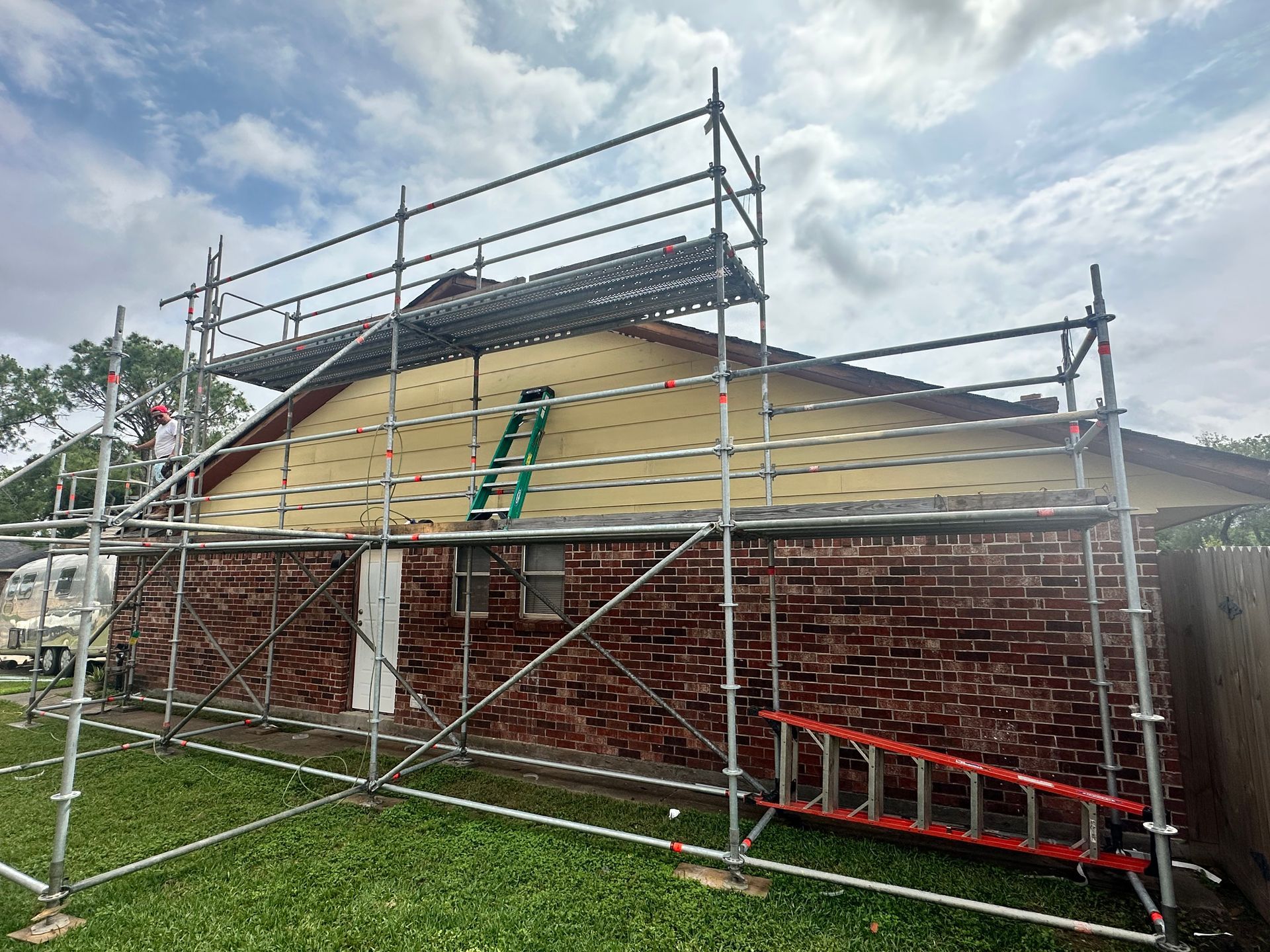 Scaffolding supports roof work on a brick house; a ladder leans against the roof.