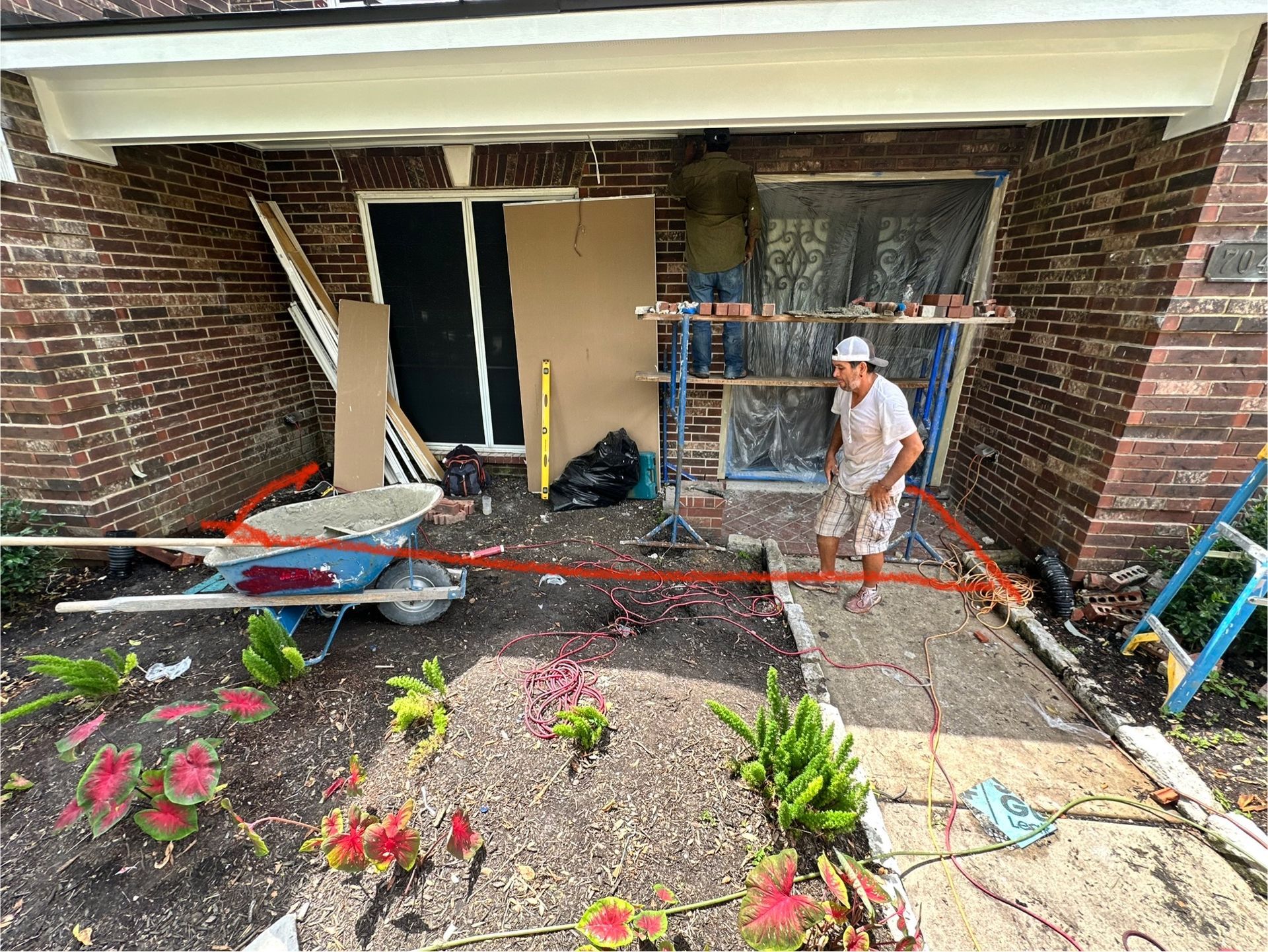 Two people renovating a brick home's entryway. Scaffolding, debris, tools, and construction materials are visible.