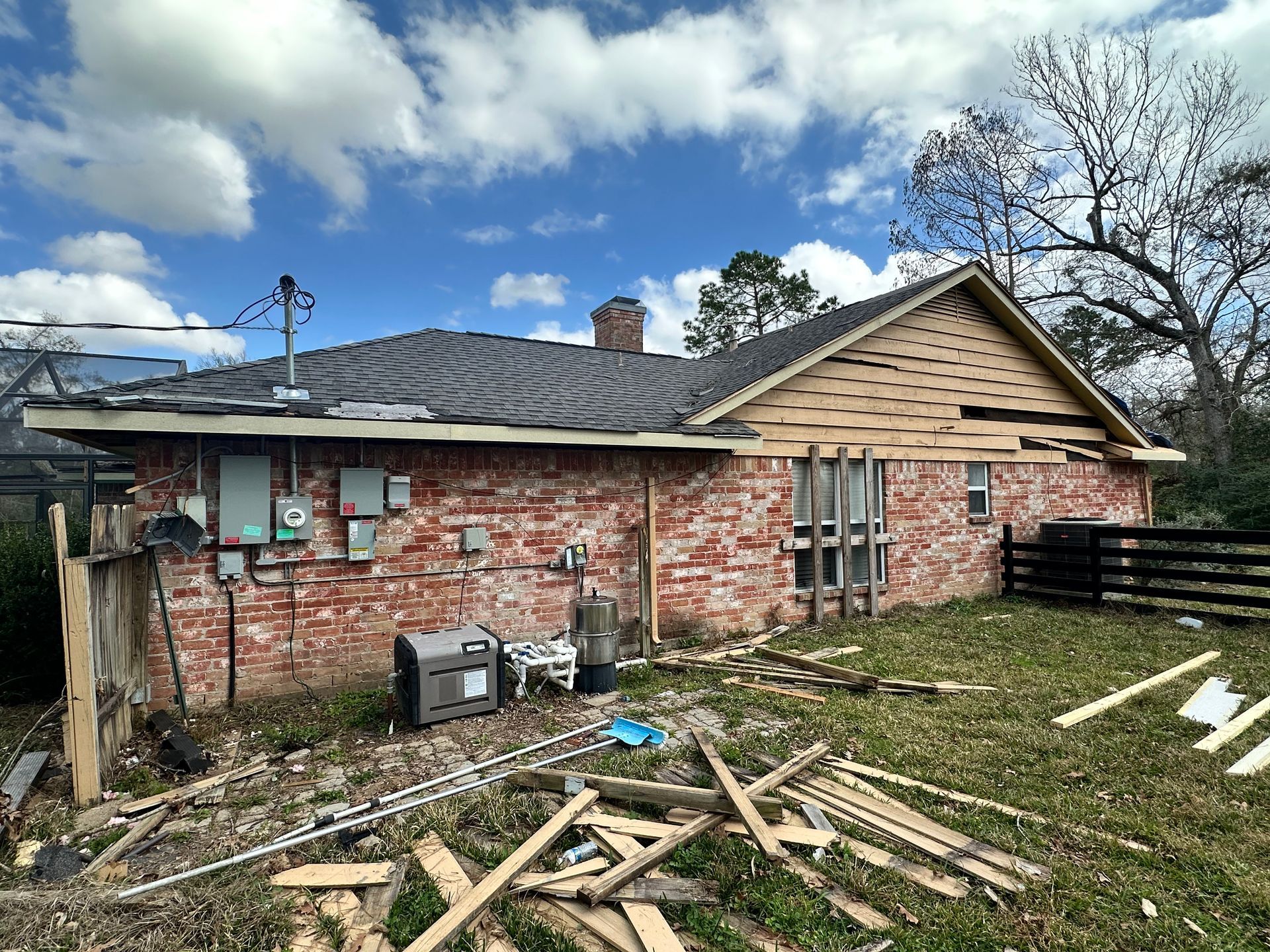 A red brick house under renovation. Roofing removed, surrounded by debris. Blue sky with clouds overhead.
