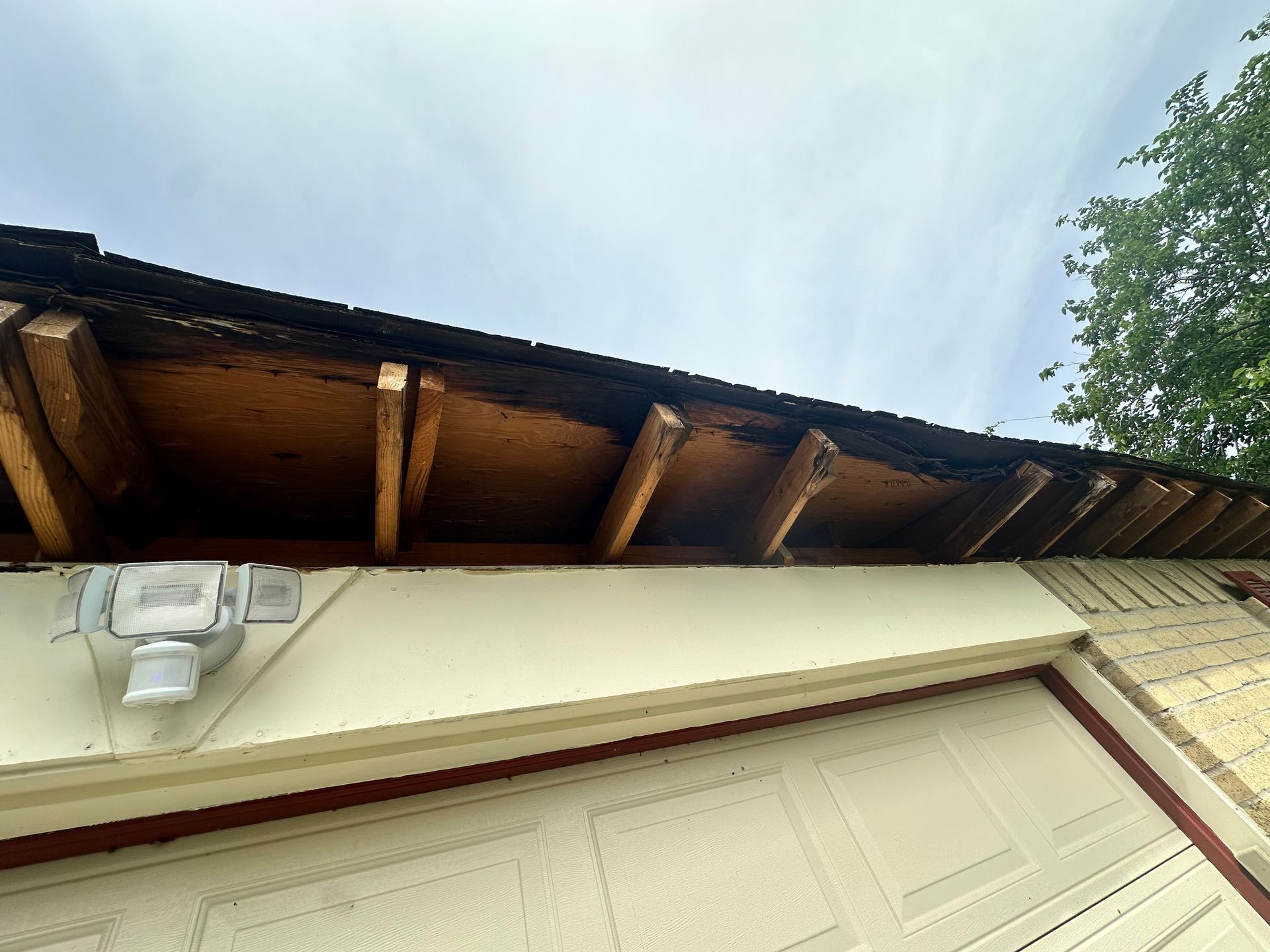 Underside of a roof overhang, showing exposed wooden supports and dark roofing material against a cloudy sky.