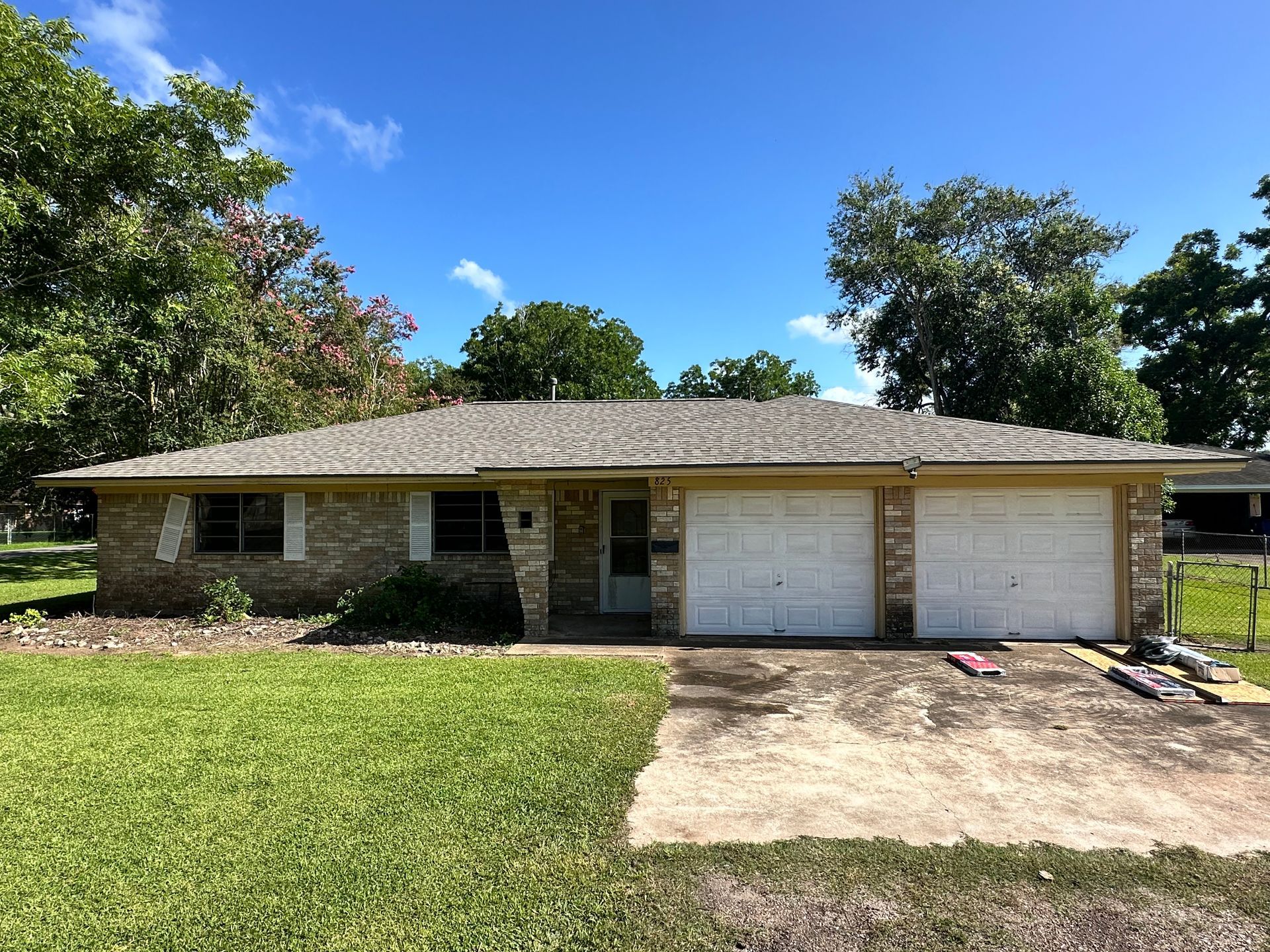 Tan brick house with a two-car garage, green lawn, and trees under a blue sky.