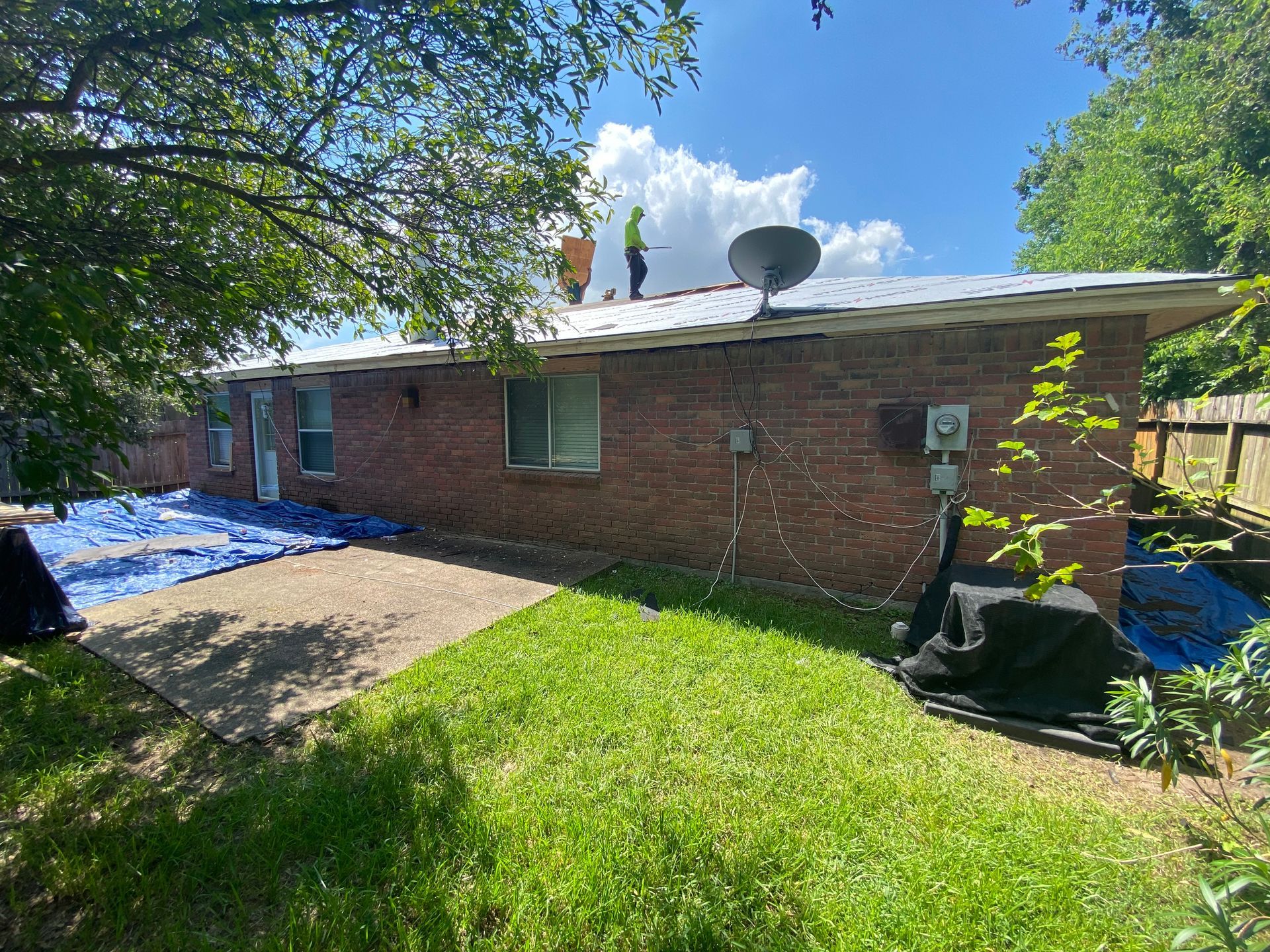 Roofers working on a brick house roof under a sunny, blue sky. Tarps cover the ground.