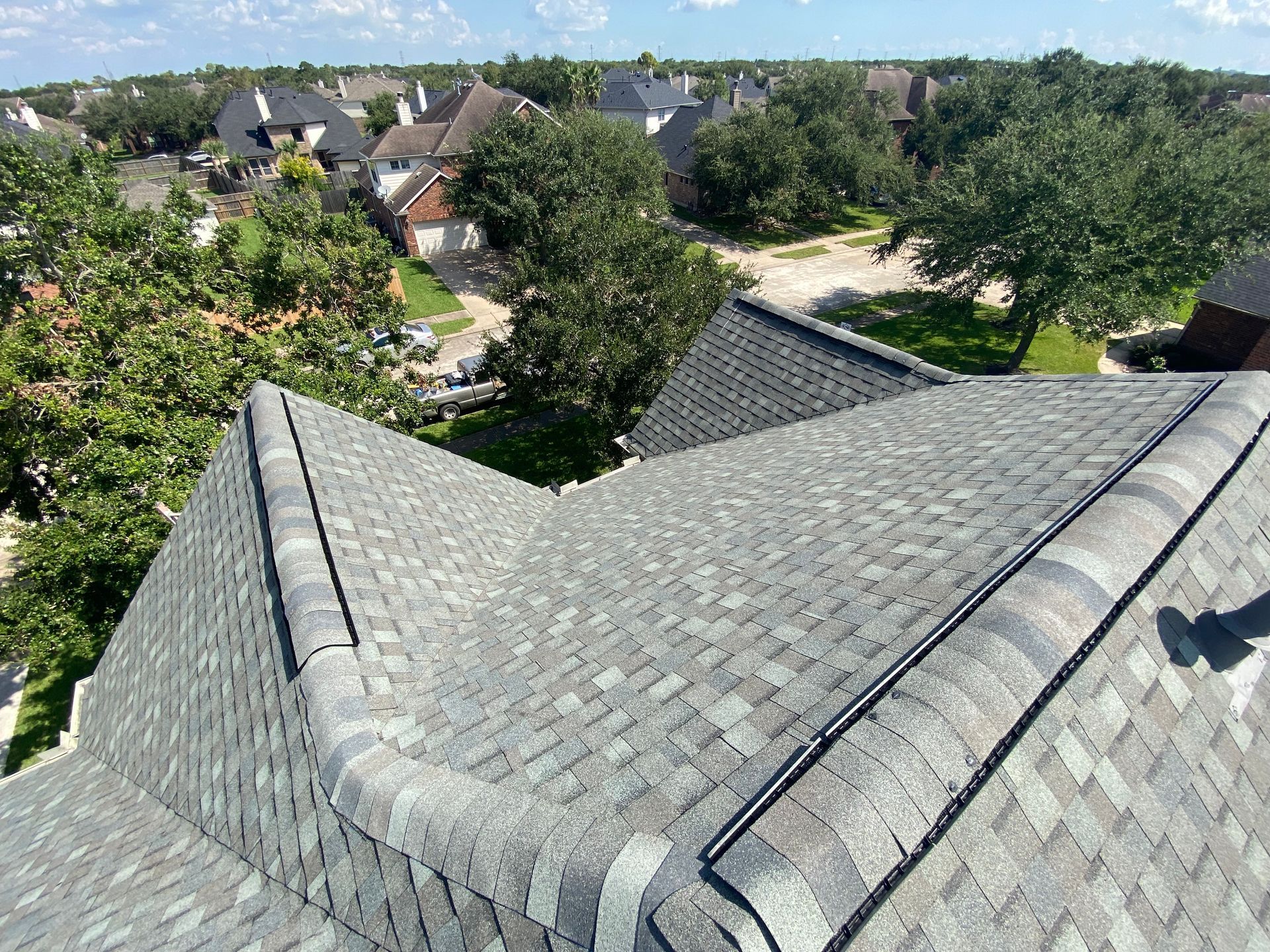 Overhead view of a gray shingled roof with a cityscape background and trees.