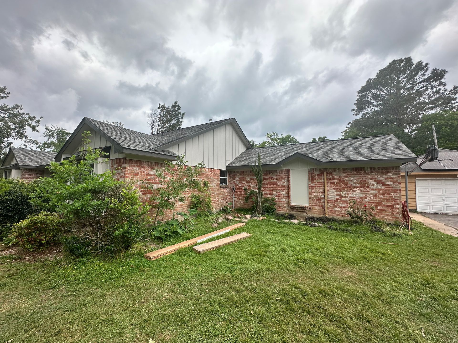 Brick house with a gray roof and overcast sky. Lush green grass and overgrown bushes.