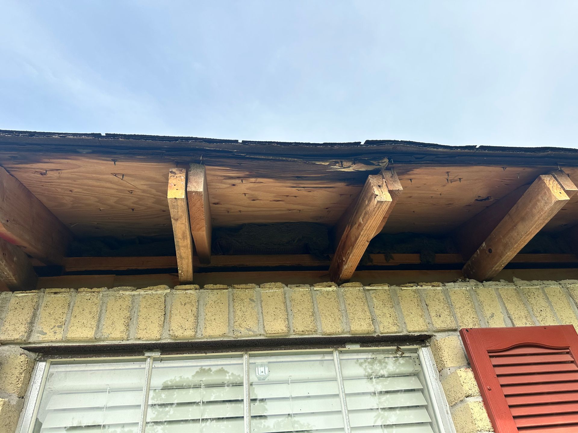 Underside of a roof overhang showing wooden structure, dark staining, and part of a brick wall and window.