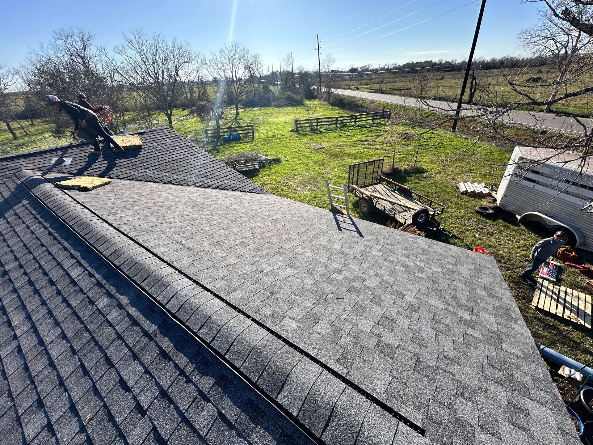 Workers installing asphalt shingles on a roof outdoors with a sunny sky and green field.