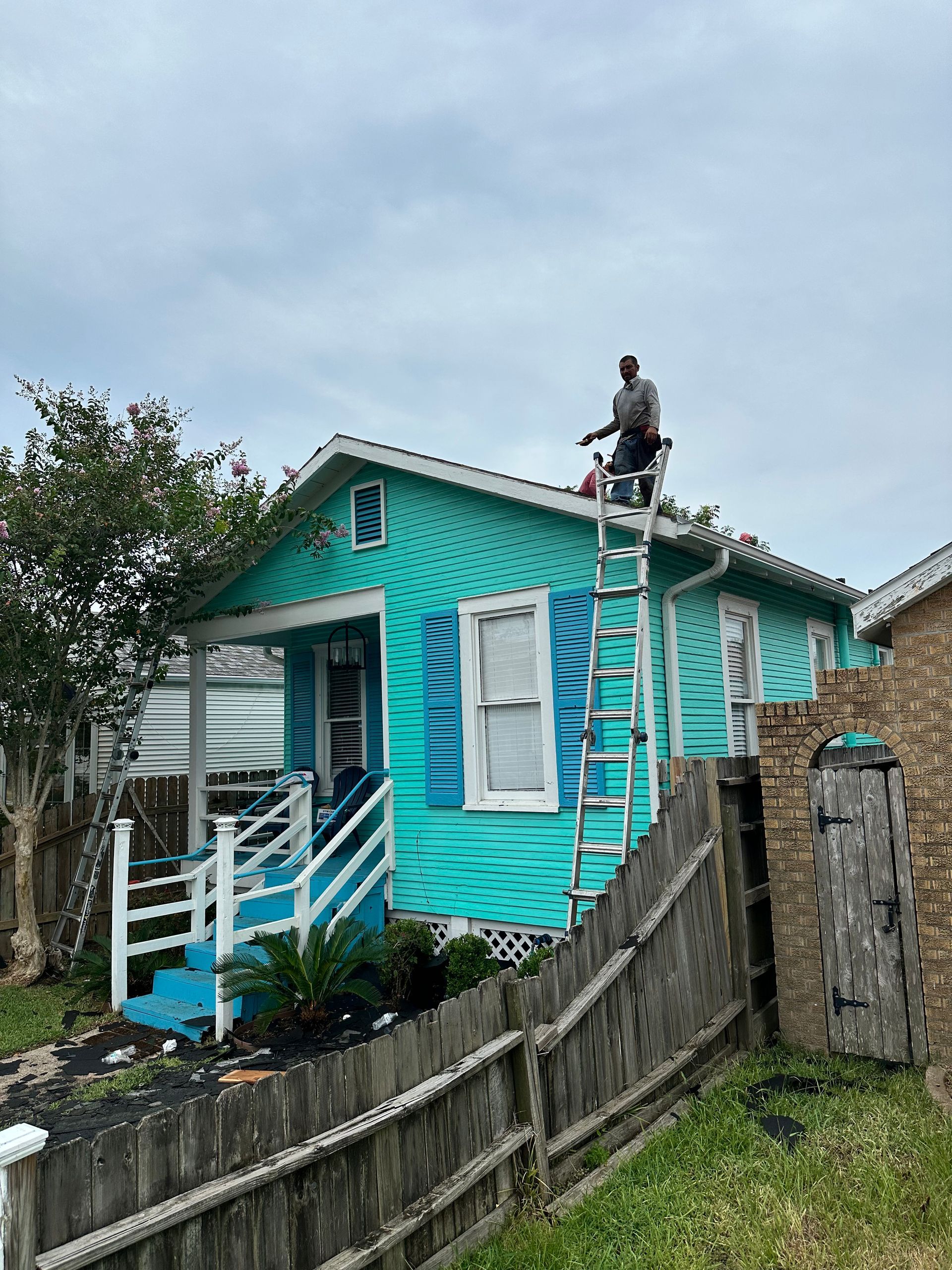 A person on a ladder working on the turquoise roof of a small house with blue shutters.