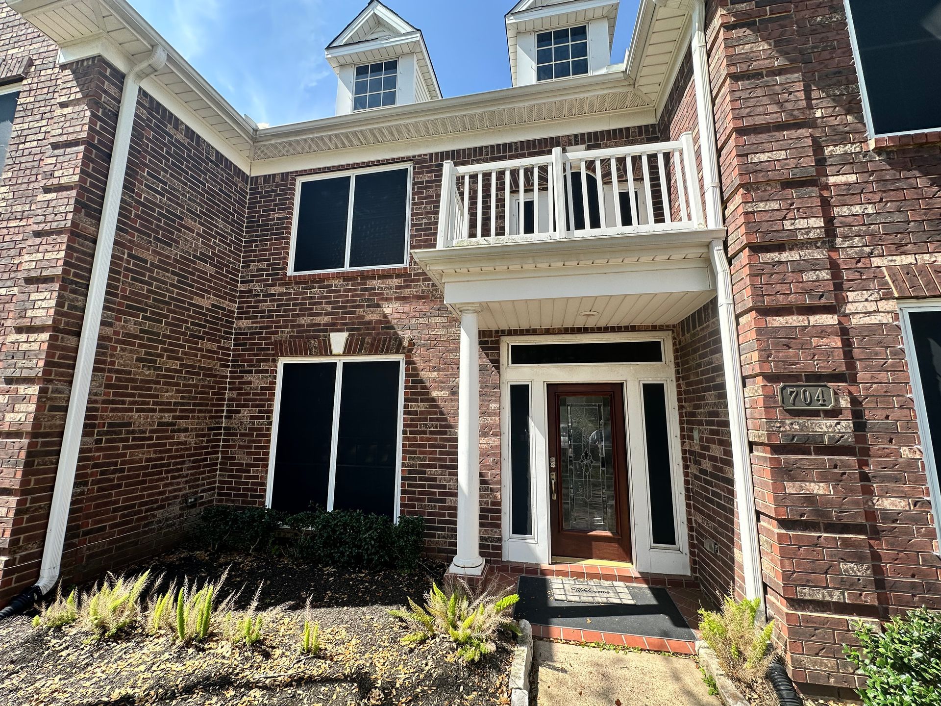 Red brick house with a white balcony and front door, featuring dark window screens.