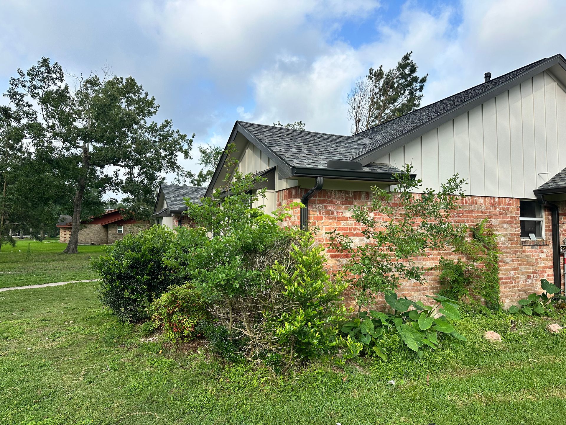 A brick and siding building, partially obscured by green bushes, under a cloudy sky.