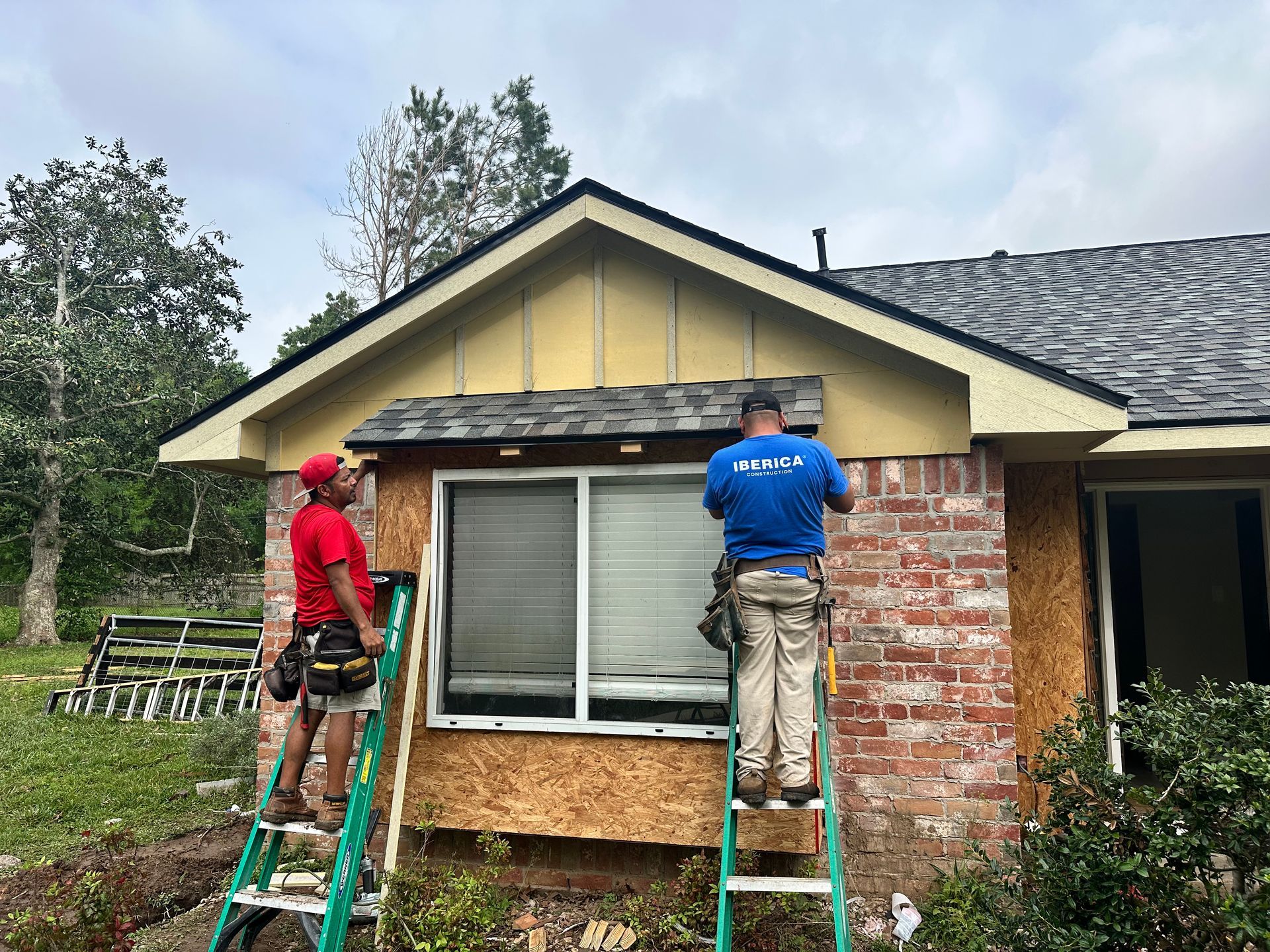 Two construction workers on ladders repair the exterior of a brick house with a window.