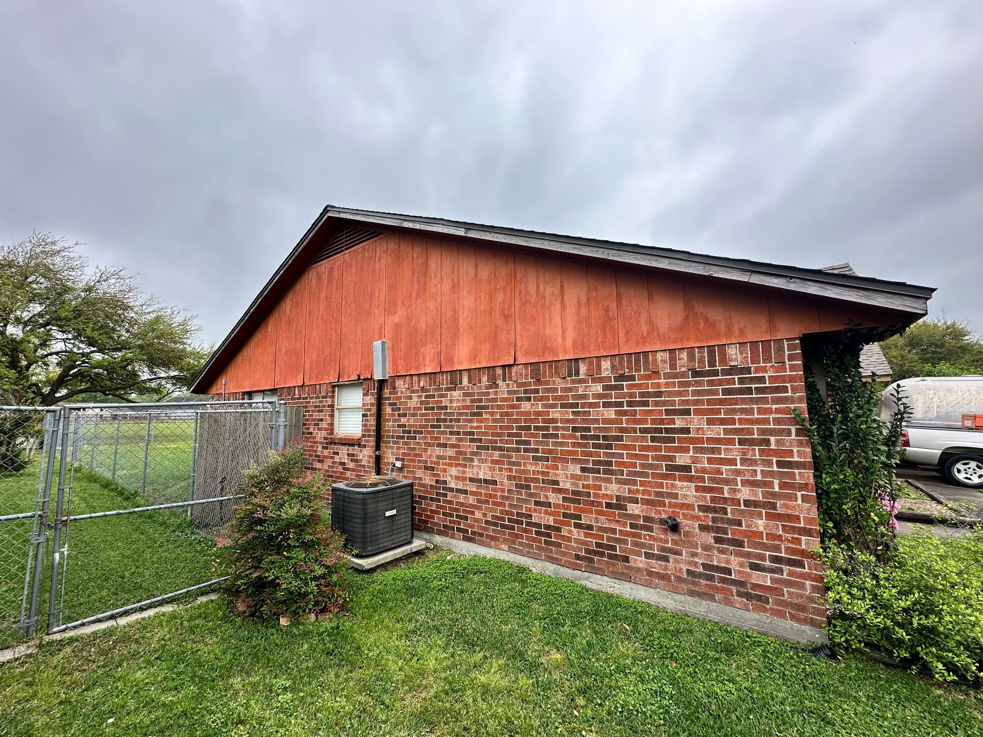 Red brick building with wooden siding, air conditioning unit, and chain link fence under cloudy sky.