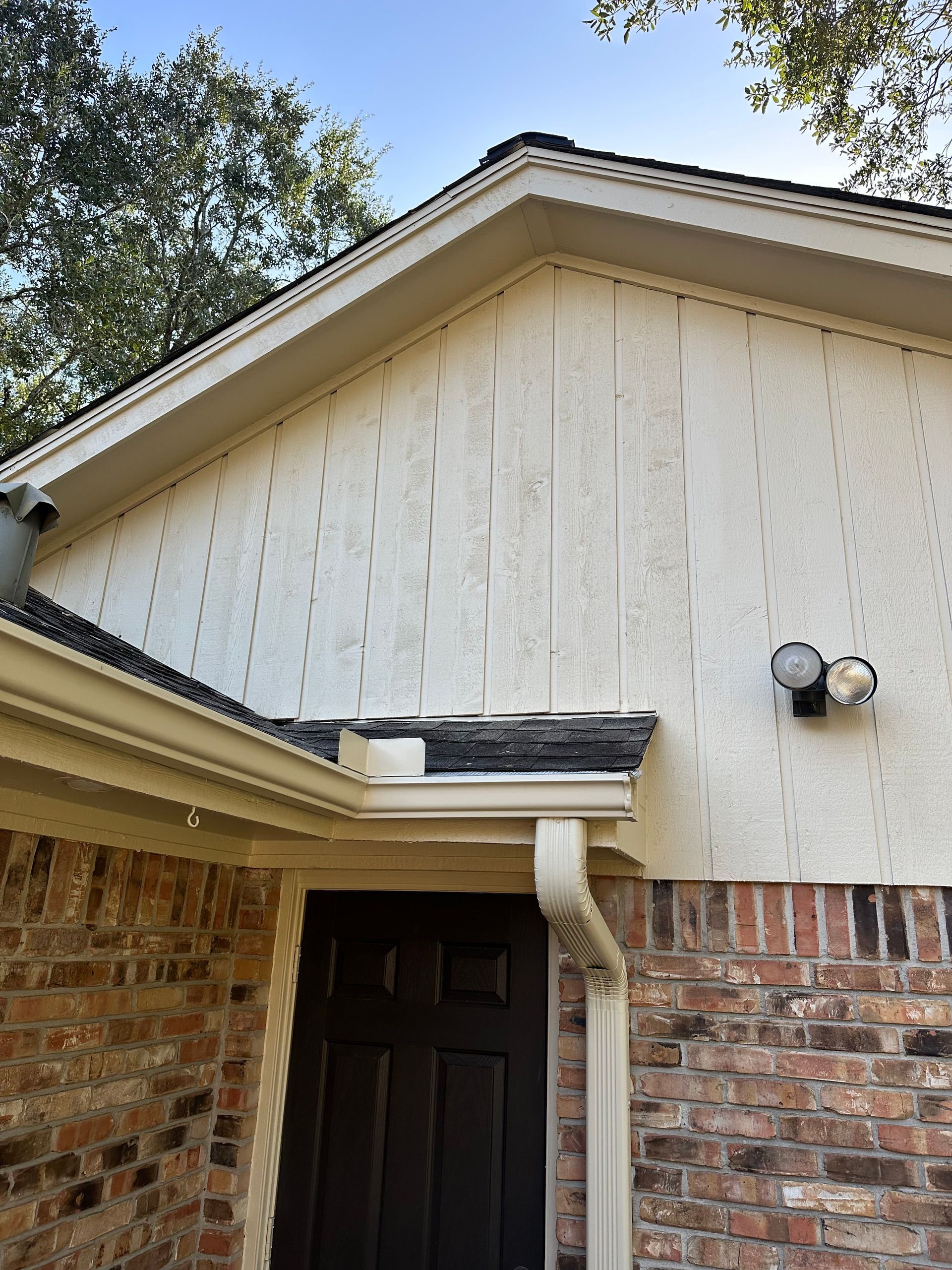 Beige siding on a house, roof, and brick exterior. White trim, gutter, and security lights.