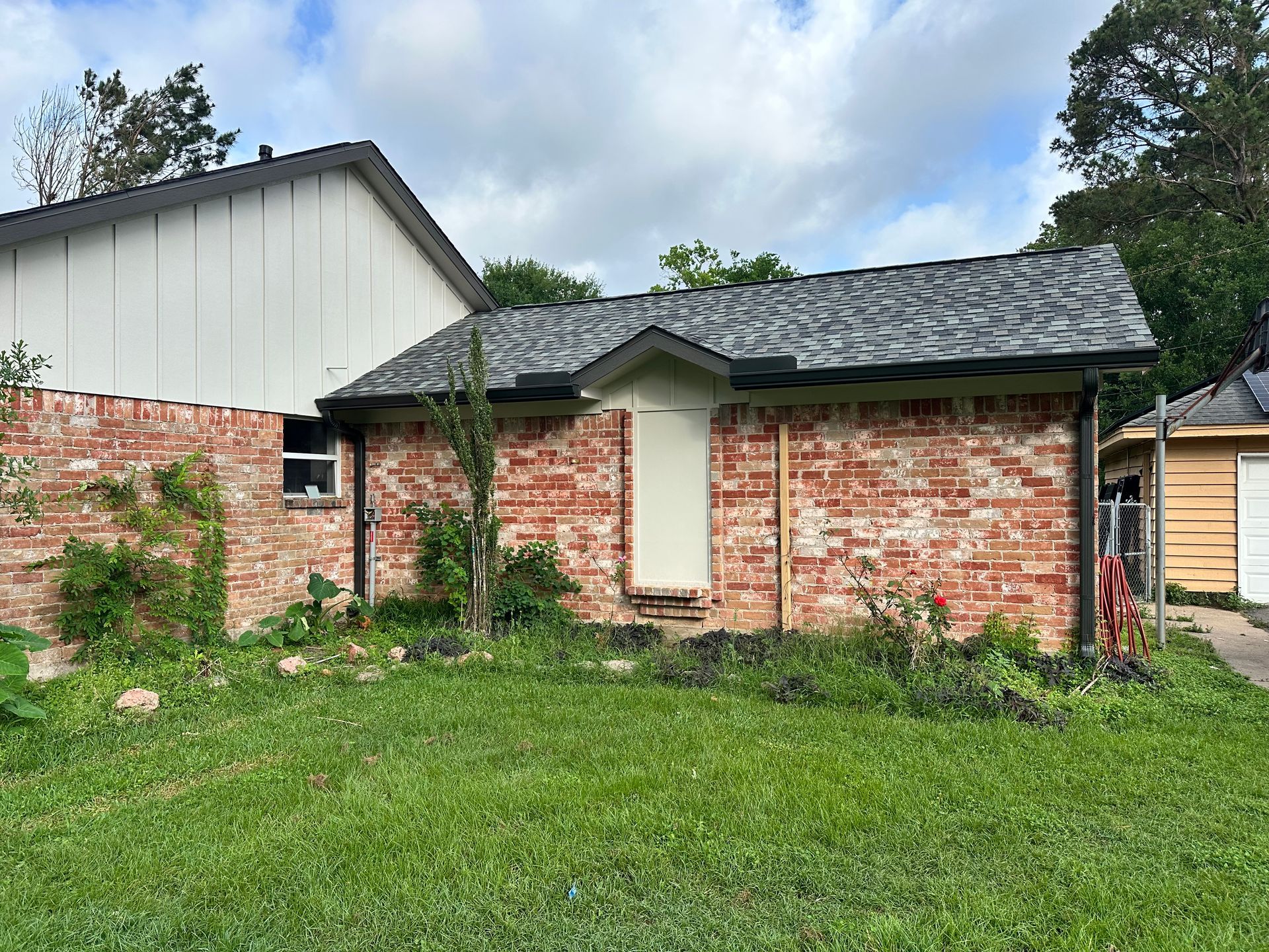 Brick building with a dark gray roof, next to a white house, on a green lawn under a cloudy sky.