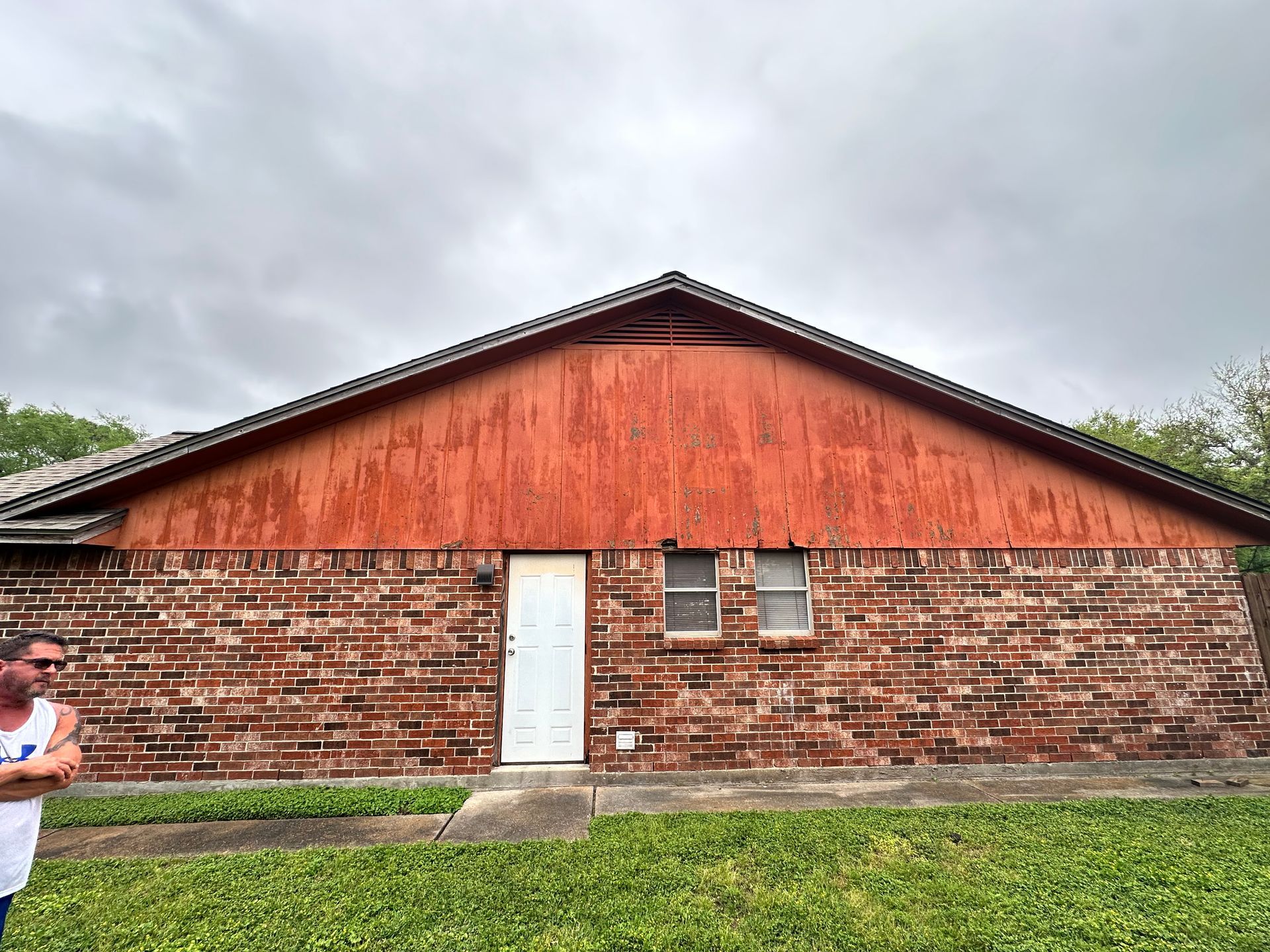 Brick building with red wood gable and door, two windows, man in tank top stands by grass.