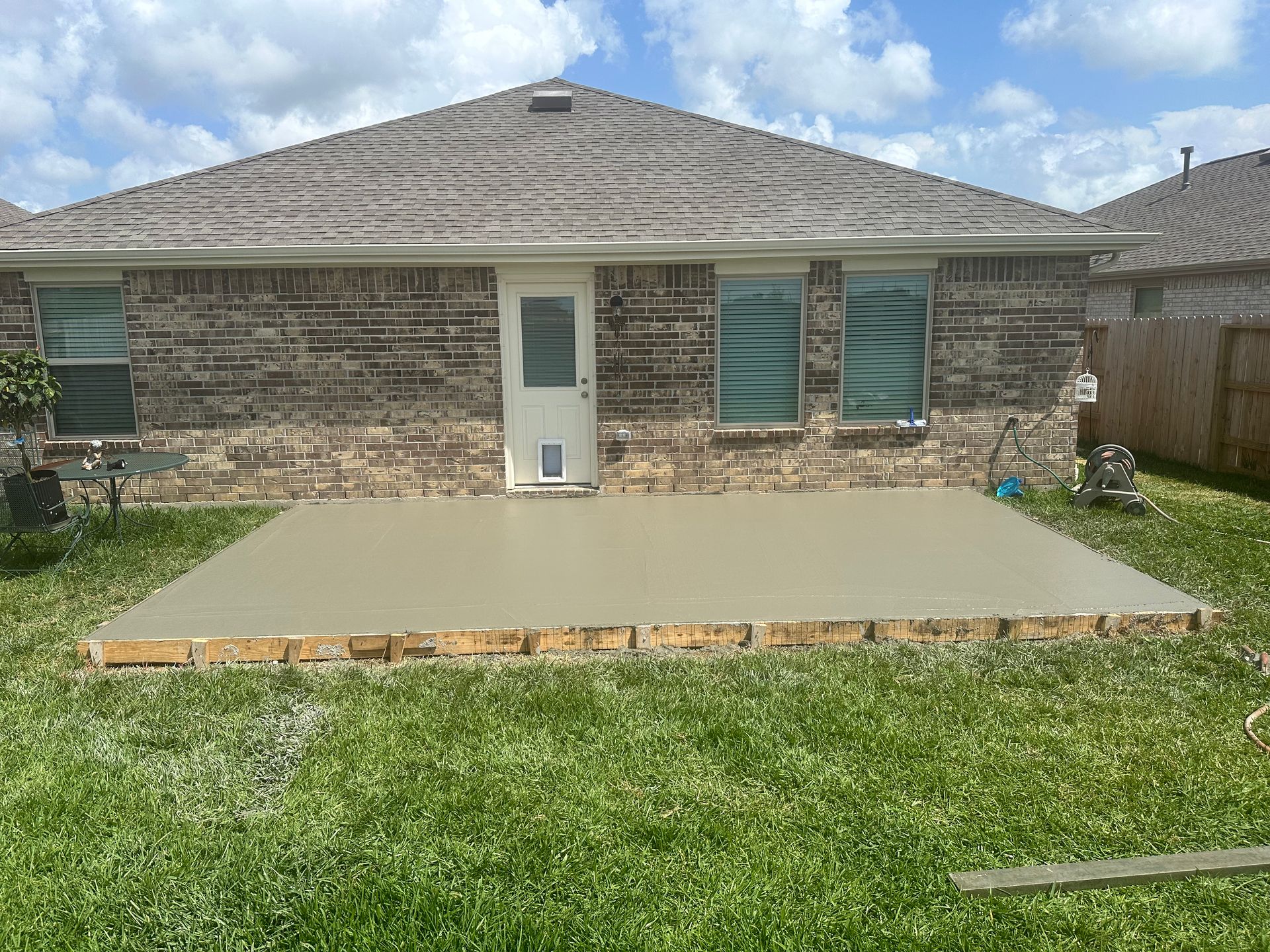 Newly poured concrete patio in a backyard against a brick house. Green grass surrounds it.