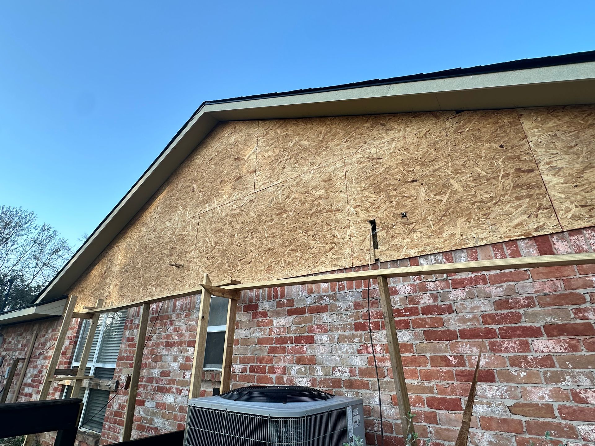 A section of a brick building with plywood siding being installed. Wooden supports prop the plywood.