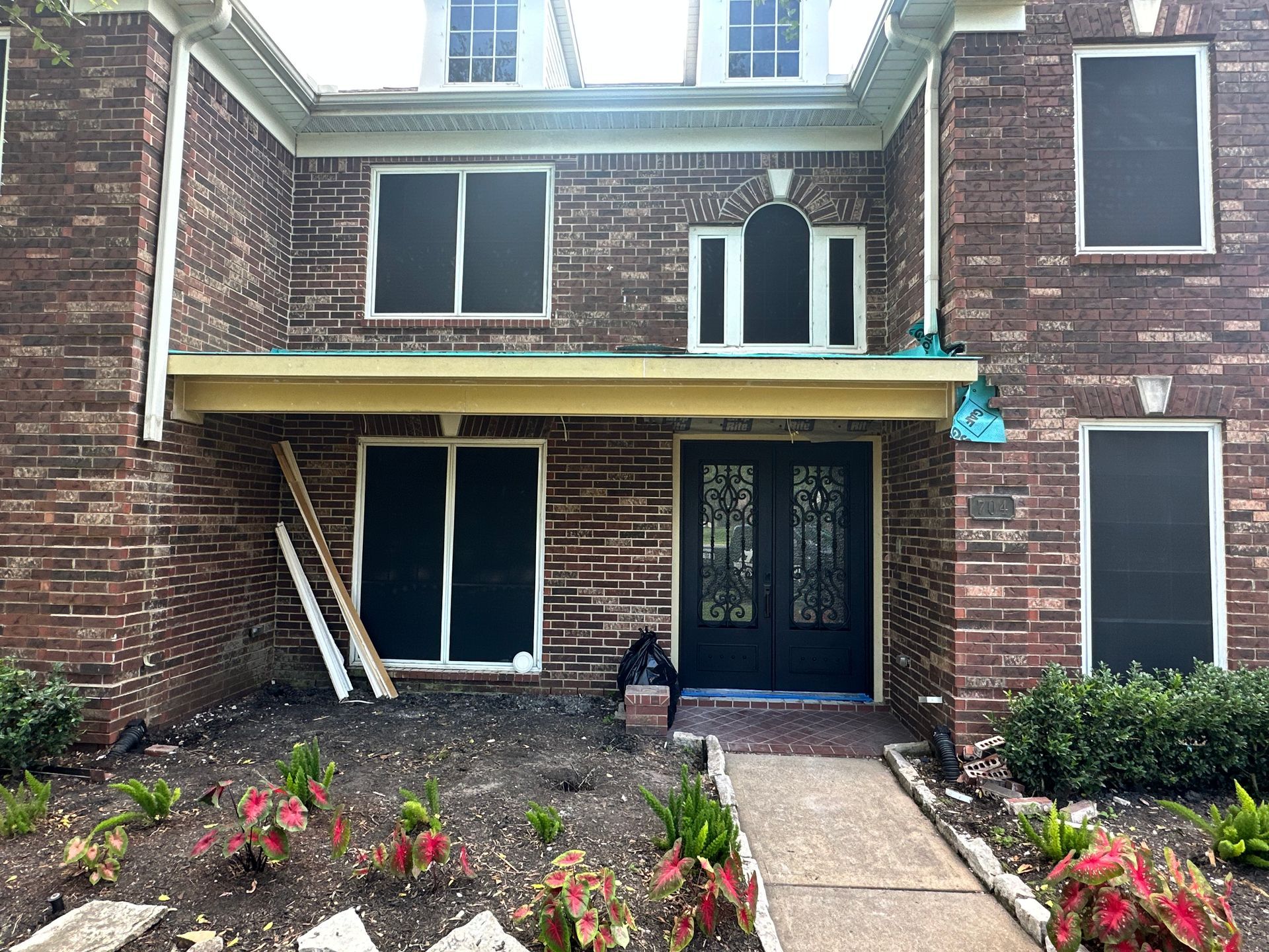 Brick house with black screen windows and double front doors, walkway, and plants.