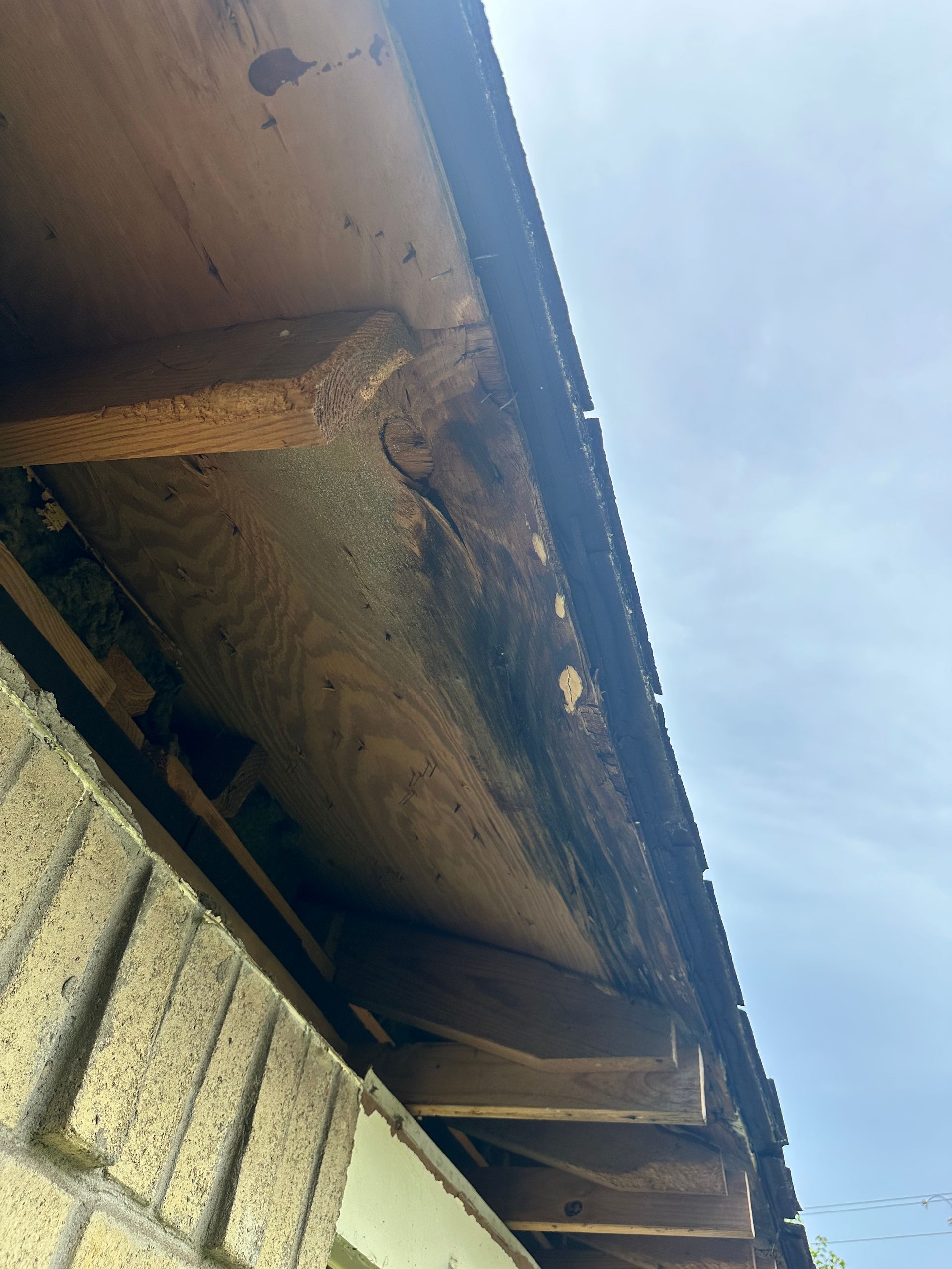 Underside of a roof's eave showing rotted wood, likely from water damage, against a partly cloudy sky.